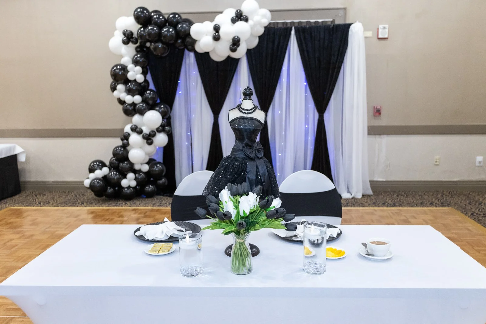 Elegant party table with black, white, and green centerpiece, black and white balloons, black and white curtains backdrop, and a black dress on a mannequin.