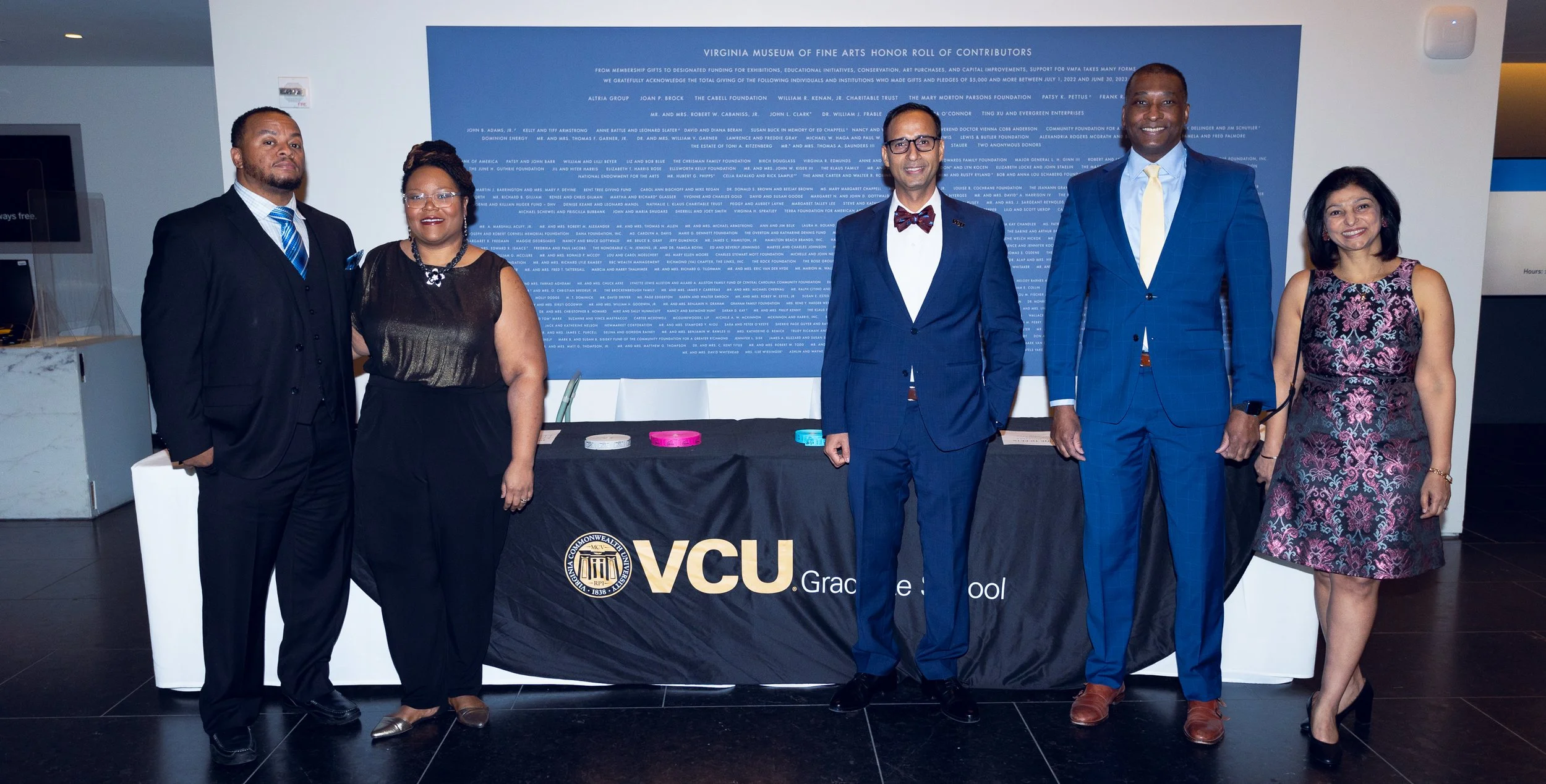 Six people standing in front of a blue informational display about the Virginia Museum of Fine Arts, with a Virginia Commonwealth University (VCU) banner on the table in front of them. They are dressed in formal attire and smiling at the camera.