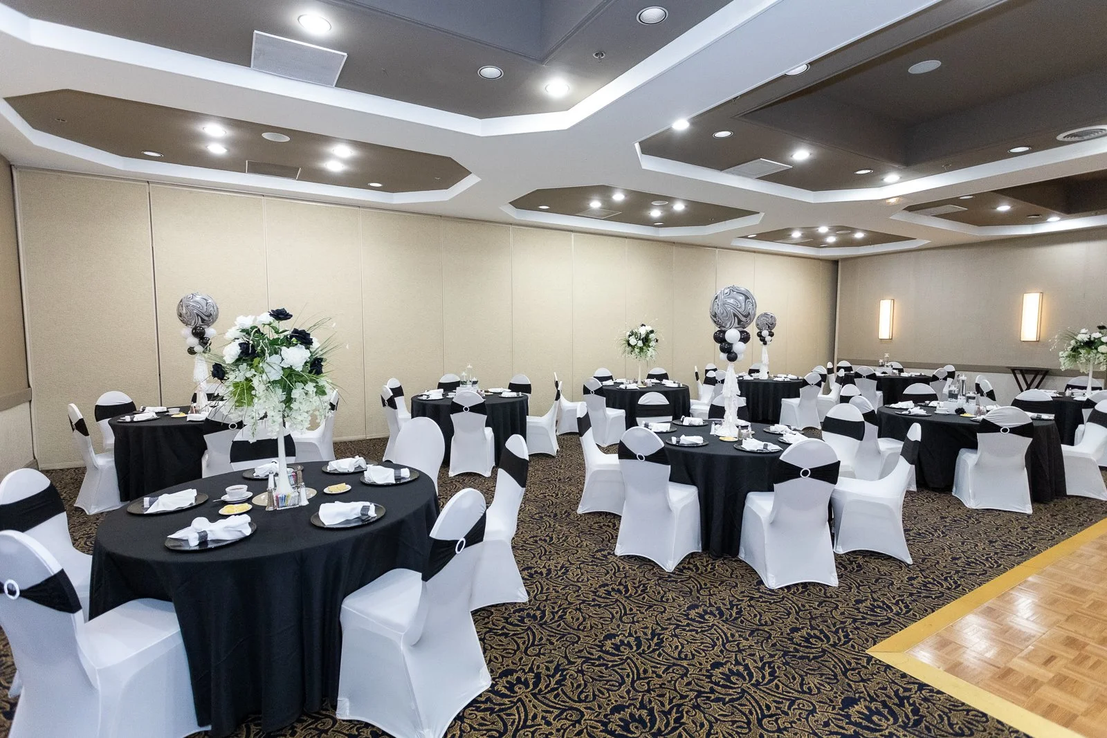 A banquet hall decorated for an event with round tables covered with black tablecloths and white chair covers with black sashes. Tall floral centerpieces and balloon arrangements in black, white, and gray are on the tables. The ceiling has modern rec