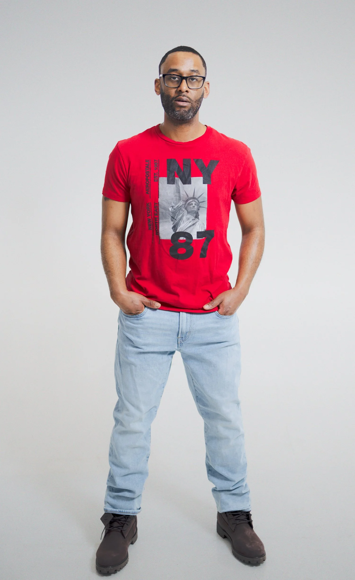 A man wearing a red T-shirt with a graphic of the Statue of Liberty and the text NYC 87, light blue jeans, and gray boots, standing against a plain white background.