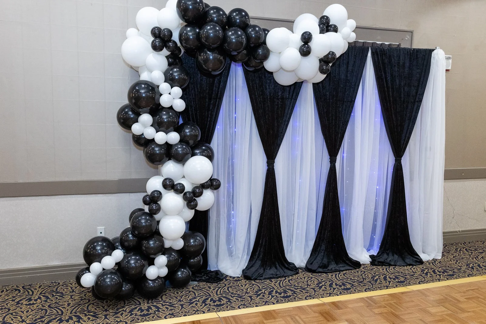 Black, white, and a few smaller balloons arranged in a balloon arch and cluster next to a black and white draped backdrop with string lights.