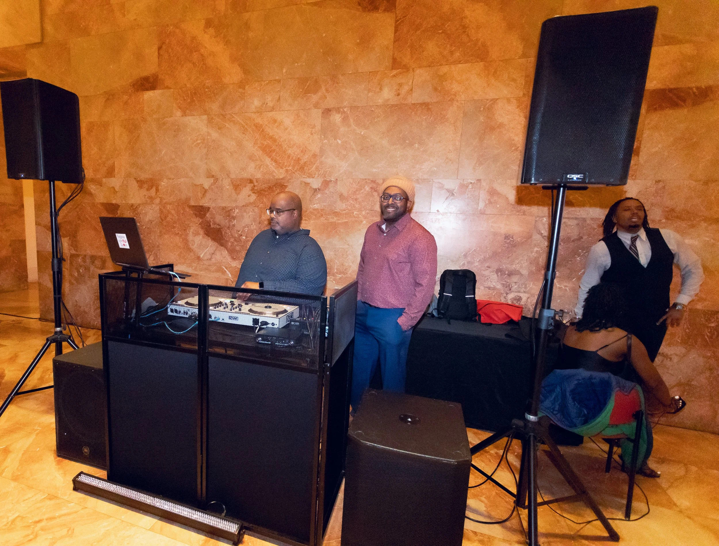 Three men are standing behind a DJ booth with equipment in a room with a marble or stone wall. One man is seated and working on a laptop, while the other two are standing, smiling and looking in different directions. Two large speakers are positioned