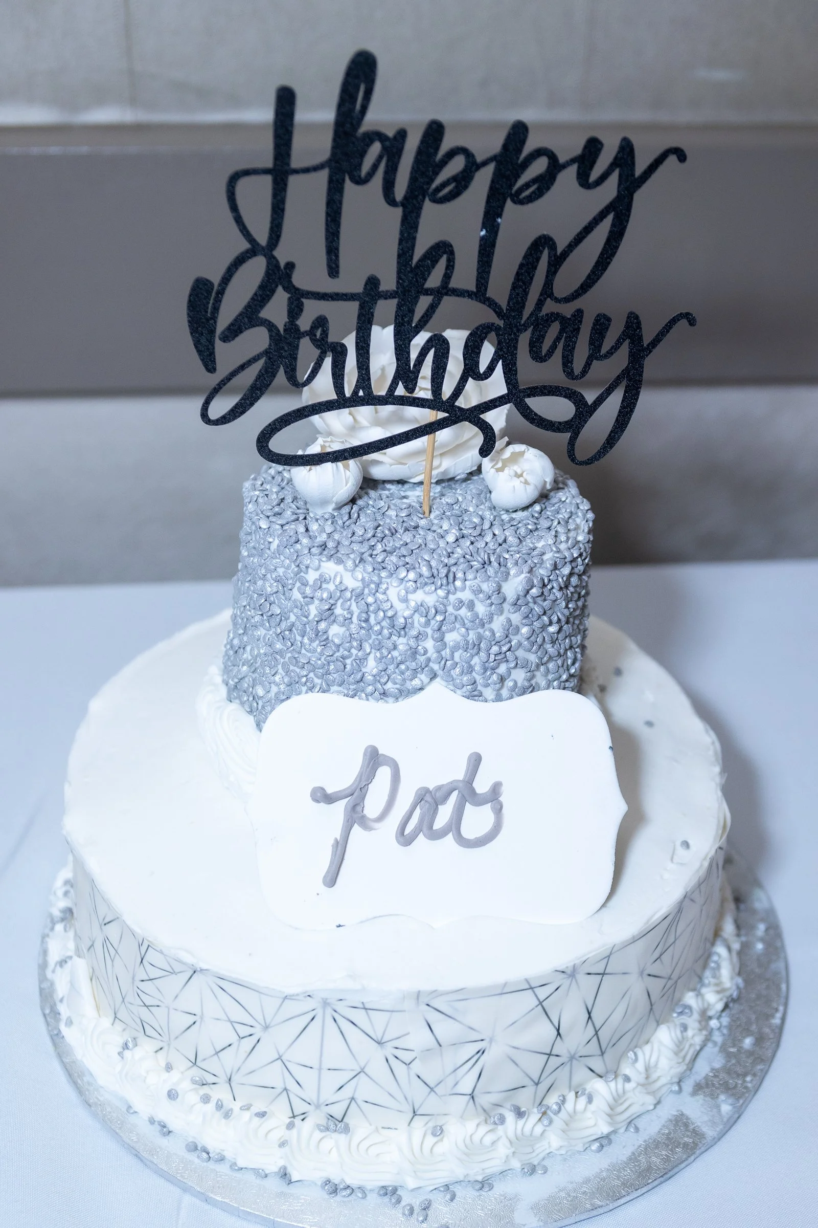 A two-tiered white birthday cake decorated with silver pearls and geometric patterns. The top tier has a silver textured surface, and the bottom tier features a delicate black geometric design. A white rectangular plaque with gray cursive writing say