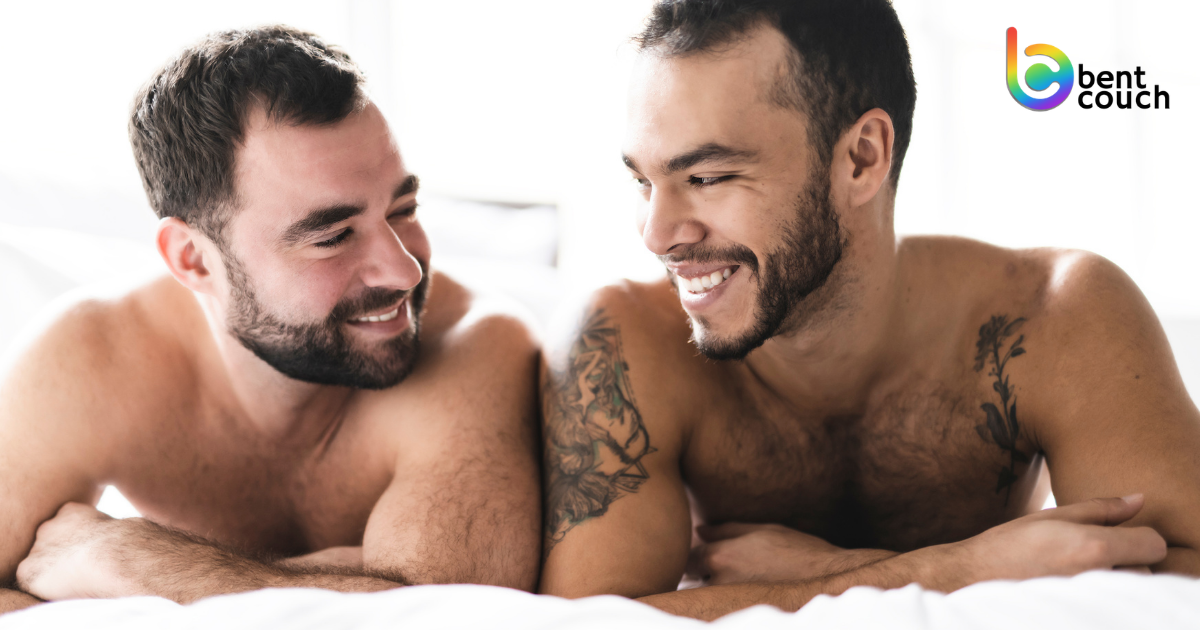 Two gay men lying close together on a bed, smiling and making eye contact, representing emotional intimacy, connection, and trust in relationships and the importance of closeness in gay men’s mental health.