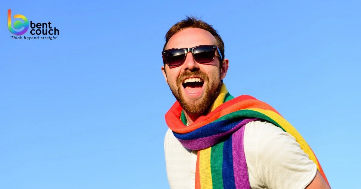 A smiling gay man wearing sunglasses and wrapped in a rainbow flag stands against a blue sky, symbolising identity, protection, self expression, and emotional survival in gay men’s mental health.