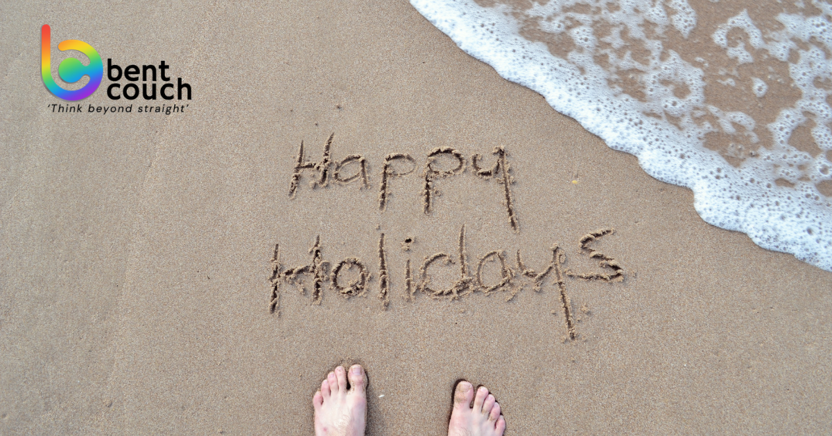 The words “Happy Holidays” written in beach sand beside the ocean, symbolising rest, time away, and the mental health benefits of taking regular holidays to prevent burnout