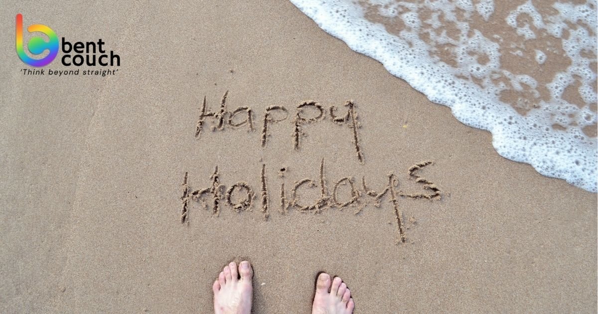 The words “Happy Holidays” written in beach sand beside the ocean, representing rest, recovery, burnout prevention, and the mental health benefits of taking regular holidays