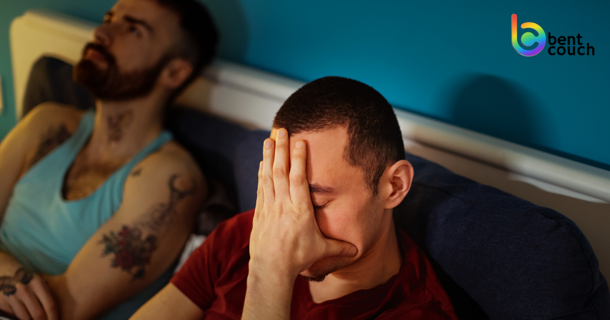 Two men sit apart on a couch, one covering his face in frustration while the other looks away, illustrating emotional distance and communication breakdown in gay couples and relationship disconnection