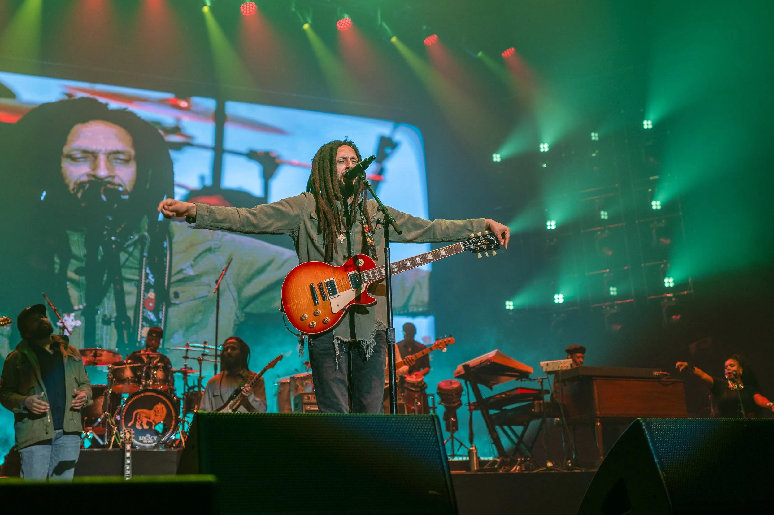 Musician with dreadlocks and a guitar performing on stage with a large video screen behind him showing his face, surrounded by other band members and colorful stage lighting.