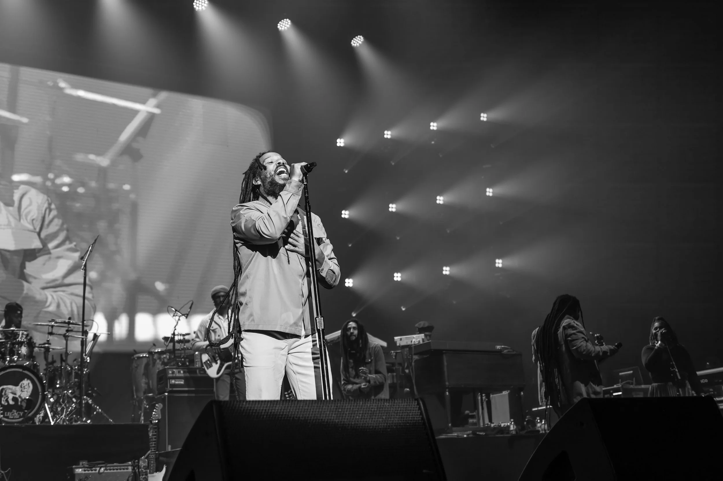 A black and white photo of a singer with dreadlocks performing on stage with a band, holding a microphone with his right hand and singing passionately. The band includes a drummer, a keyboardist, and other musicians, with stage lights shining above.