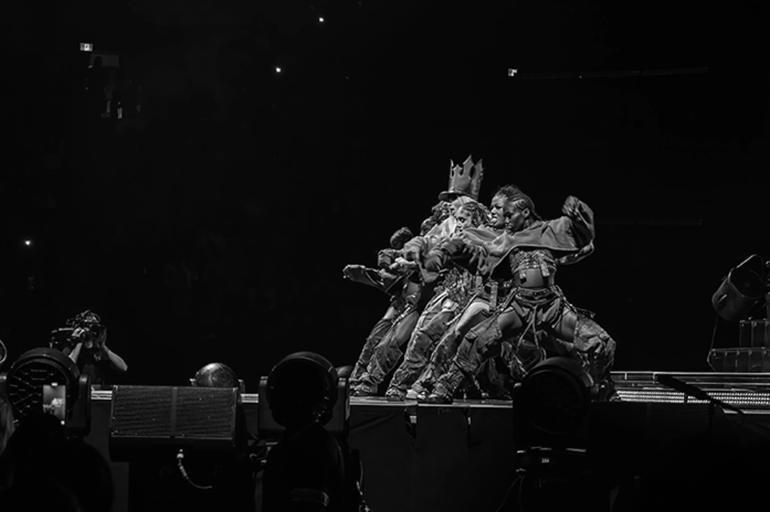 Black and white photo of dancers performing on stage with one person wearing a crown, all in military-style pants, in front of a dark background with stage lights.