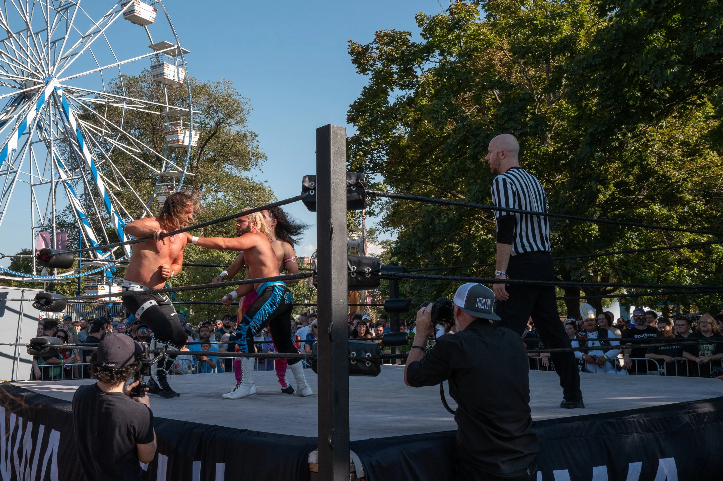 Wrestlers in a ring engaged in a match, with a referee observing and photographers capturing the event, and a ferris wheel and crowd visible in the background on a sunny day.