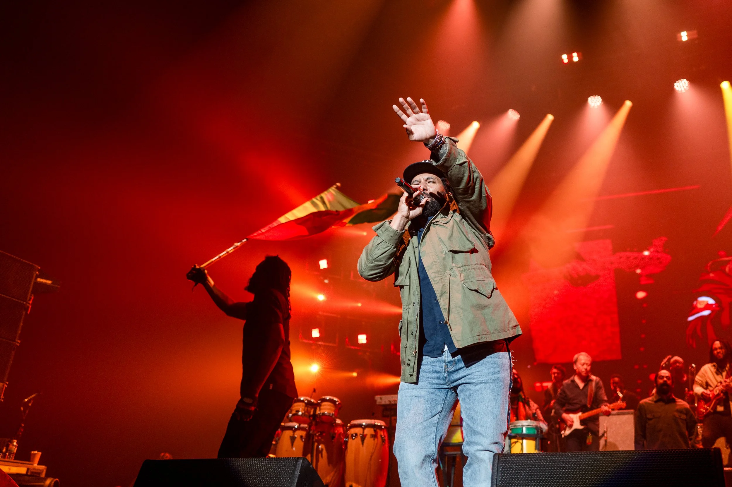 A man singing into a microphone on stage, wearing a green jacket and blue jeans, with a person in the background holding a flag. There are musicians and musical instruments, including drums and guitars, under colorful stage lights.