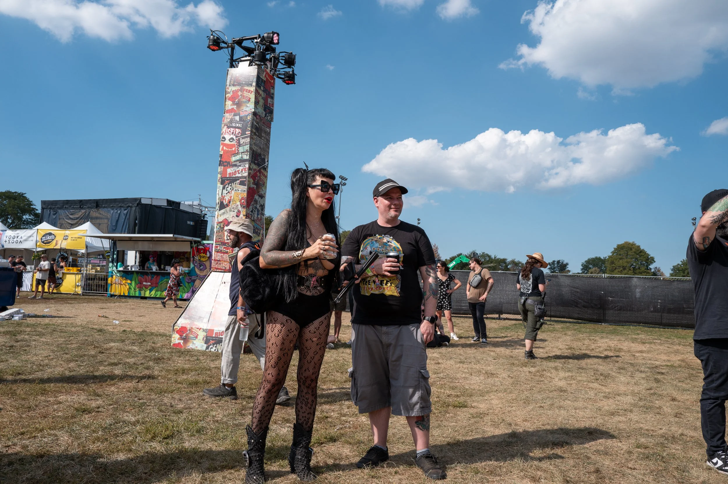 Two people at a festival, a woman with long black hair, dressed in black with fishnet stockings and sunglasses, holding a drink, and a man wearing a black t-shirt, shorts, and a cap, also holding a drink. Background includes festival tents, a tall de