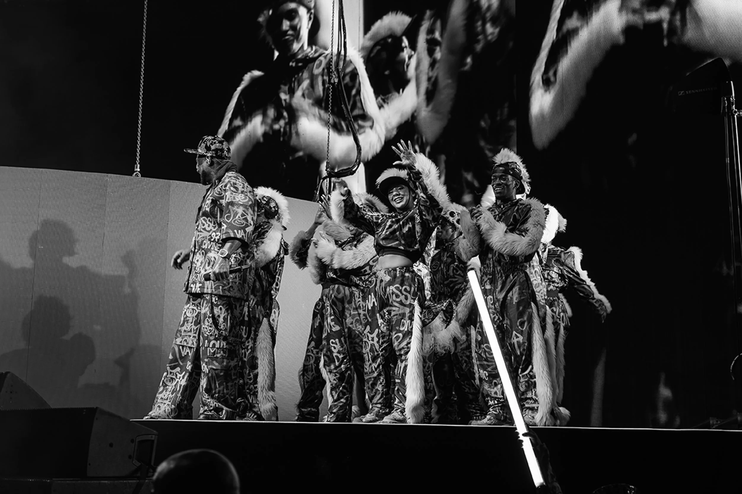 A group of performers on stage dressed in patterned outfits with fur accents, some wearing feathered headdresses, one of whom is smiling and waving. A large screen behind them displays an enlarged image of the group.