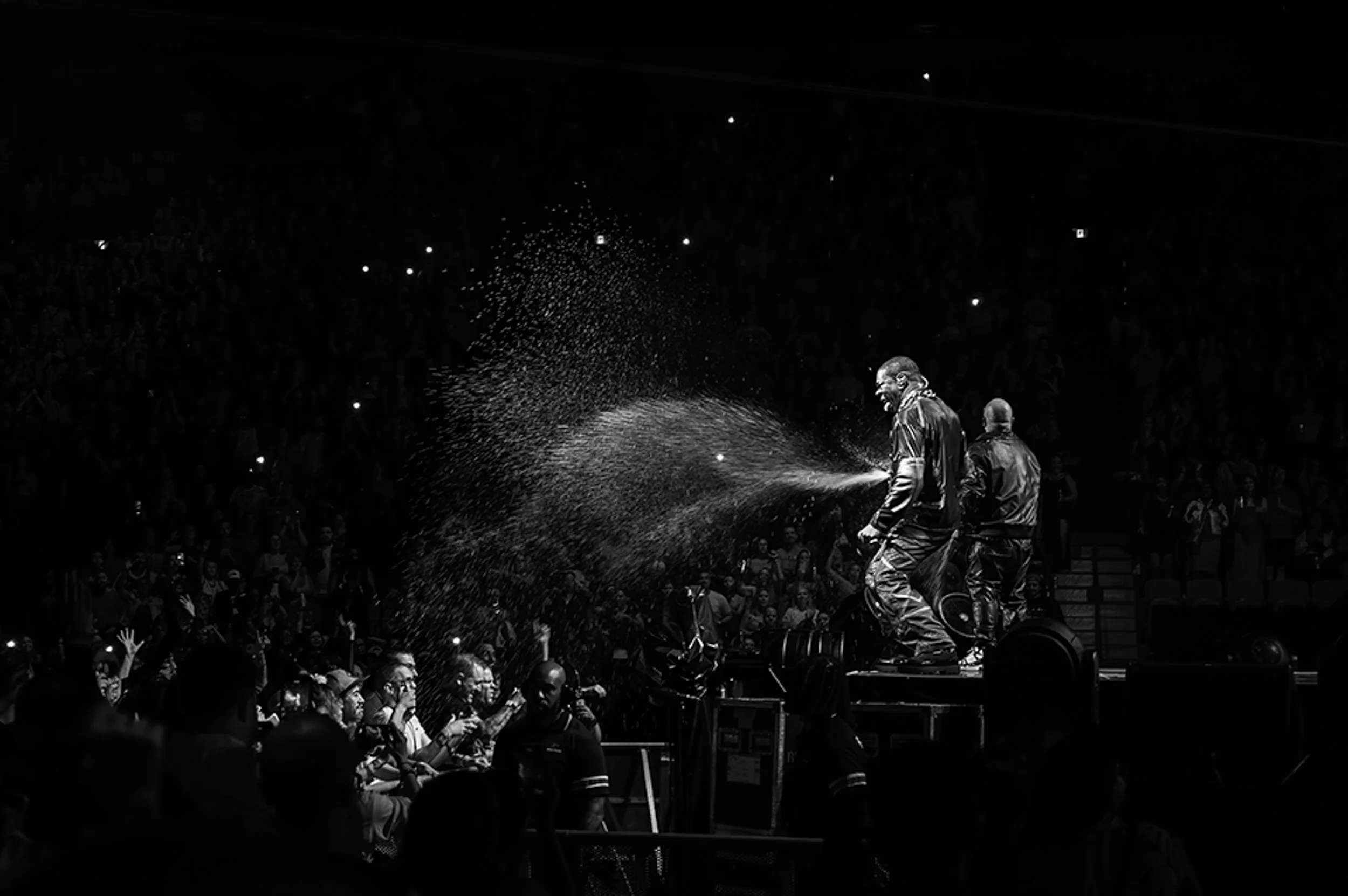 Black and white photo of two performers standing on a stage, spraying water towards the audience during a concert or event, with audience members raising their hands and cheering.
