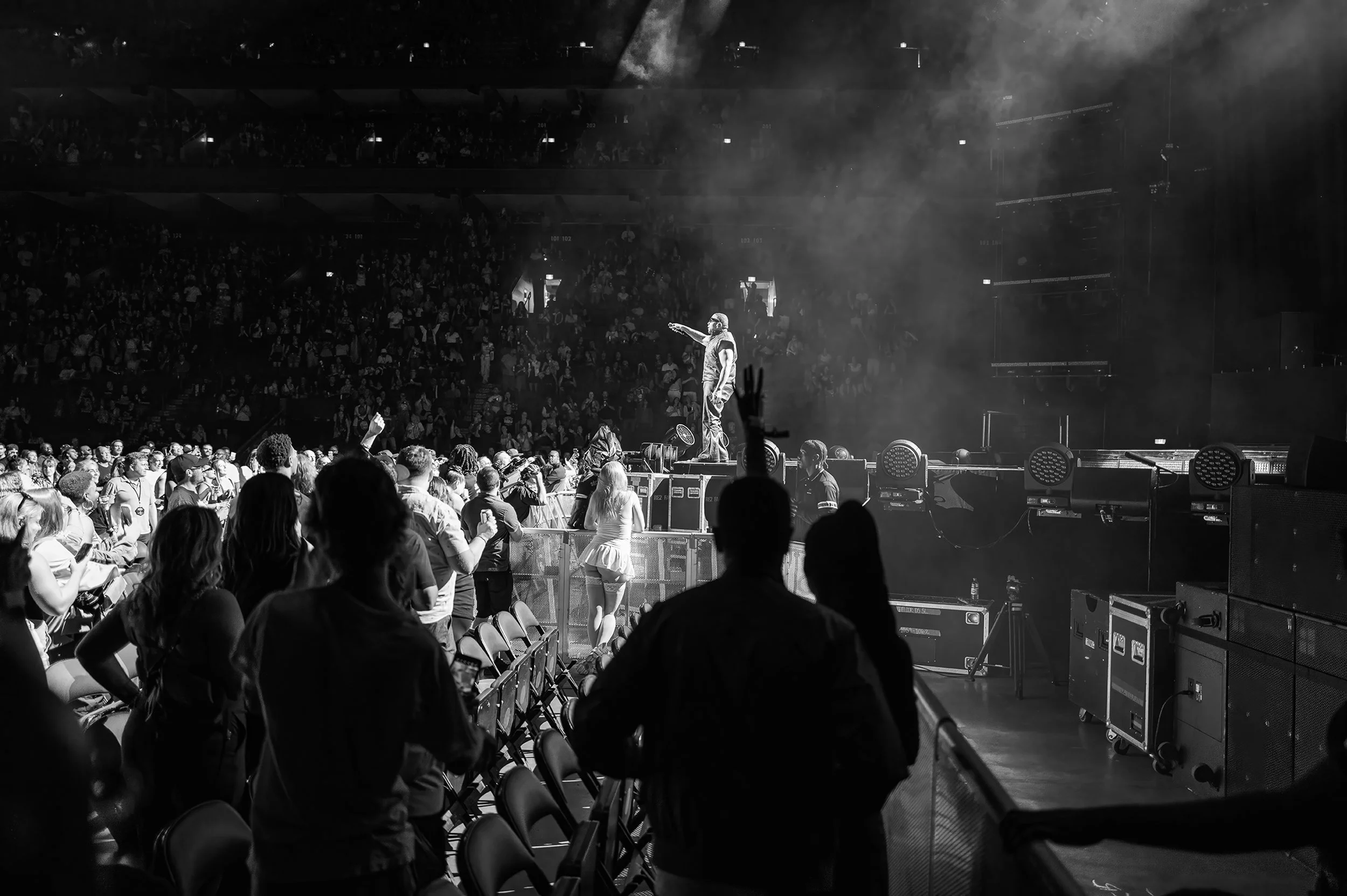 Black and white photo of a concert with a performer on stage, audience in front, and seats filled in the background. Some people are standing, watching, and taking pictures.