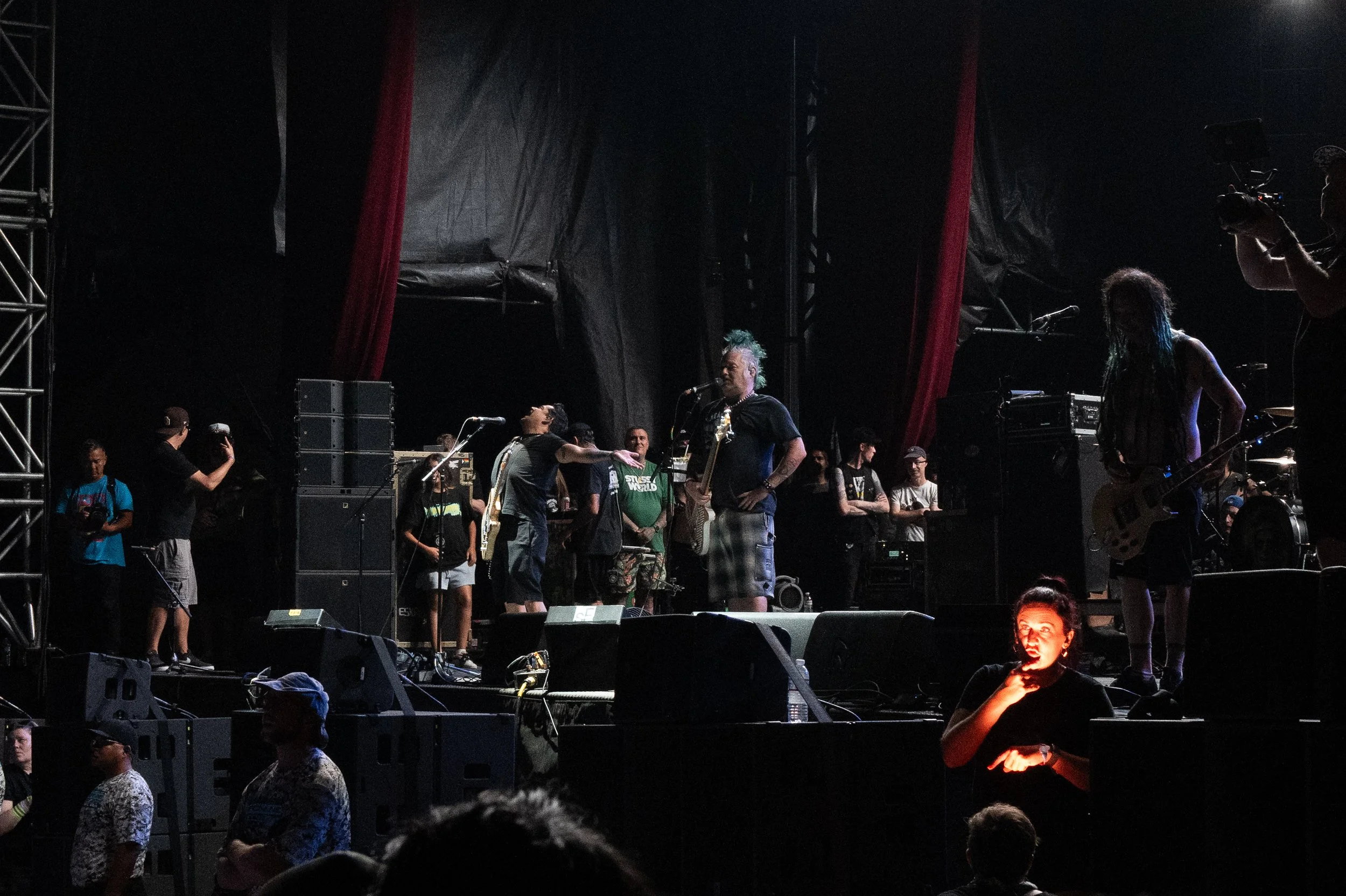 Musicians preparing for a concert on stage, with sound equipment and crew members backstage, in a dark setting.