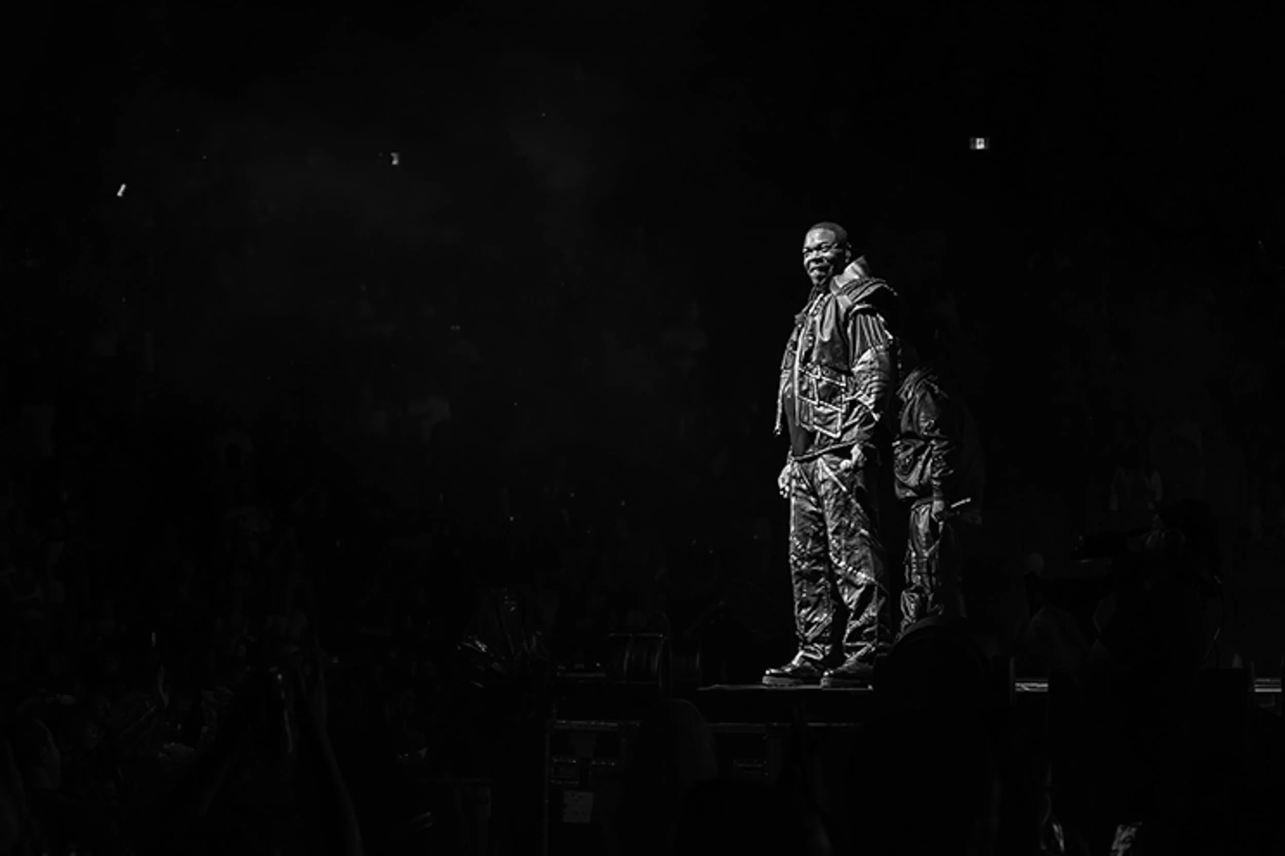 A man stands on stage, illuminated by spotlight, with an otherwise dark background. He is dressed in camouflage pants and a patterned shirt, smiling.