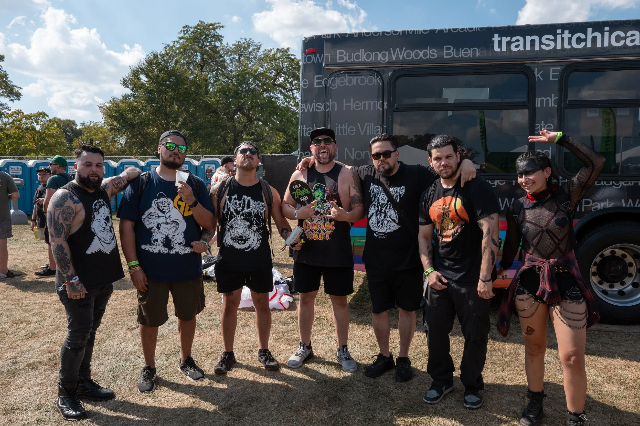 Group of seven people at a music festival standing in front of a black bus, some making playful gestures. They are dressed in casual and alternative fashion, with tattoos, sunglasses, and accessories, outdoors on a sunny day with trees and portable t