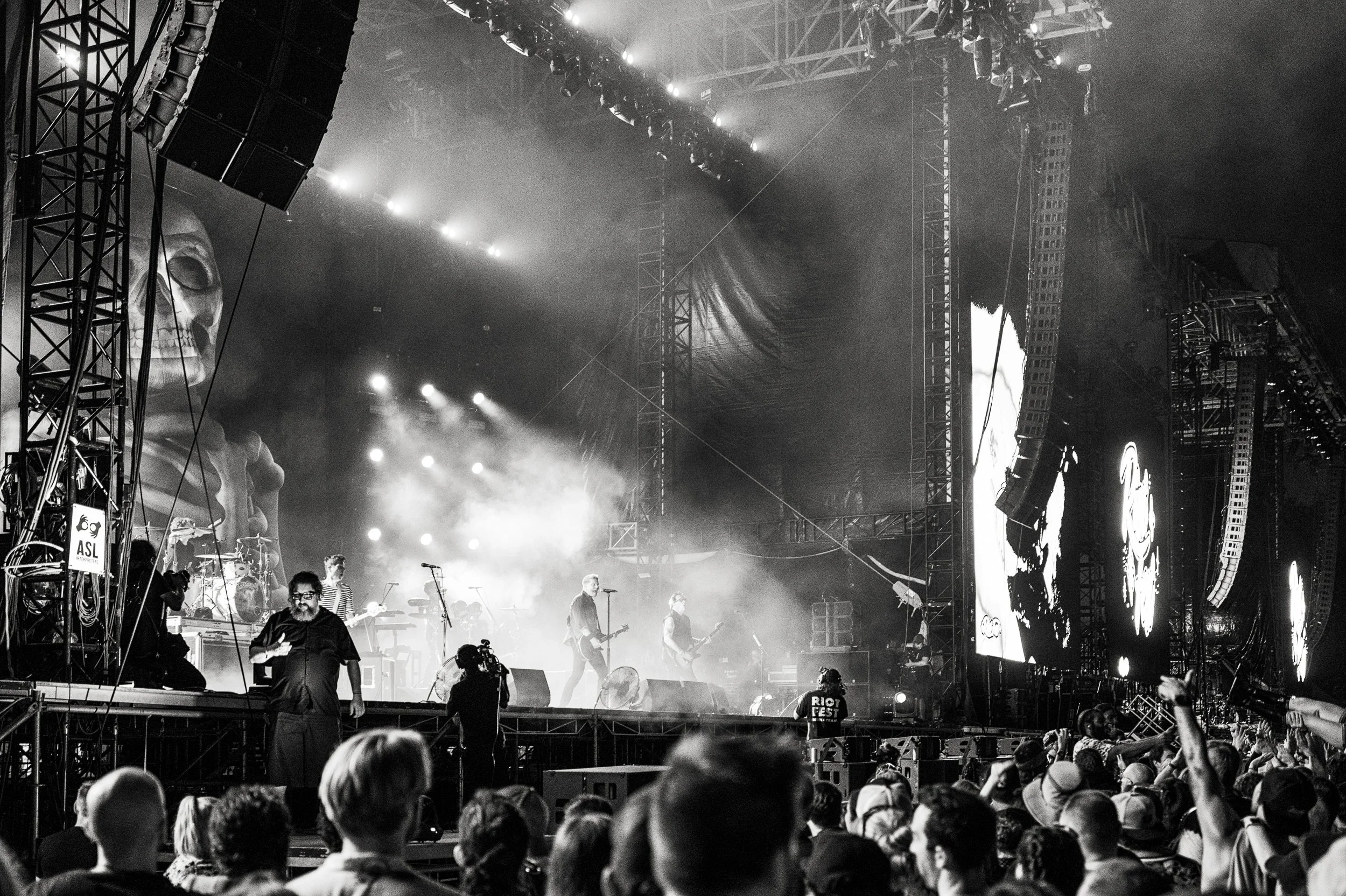 Black and white photo of a large crowd at a concert with a band performing on stage, featuring large screens and stage lights.