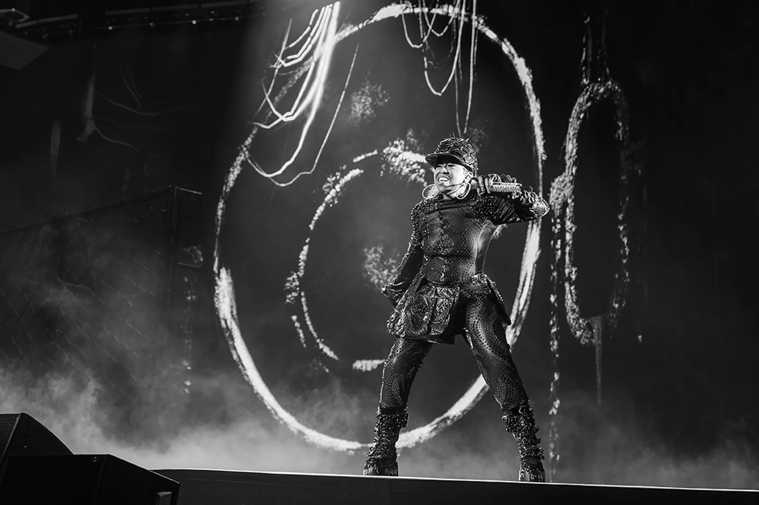 A performer dressed in a dark, shiny, futuristic costume on stage, smiling with a dynamic pose, with abstract circular graphics on a large screen behind them in a concert setting.