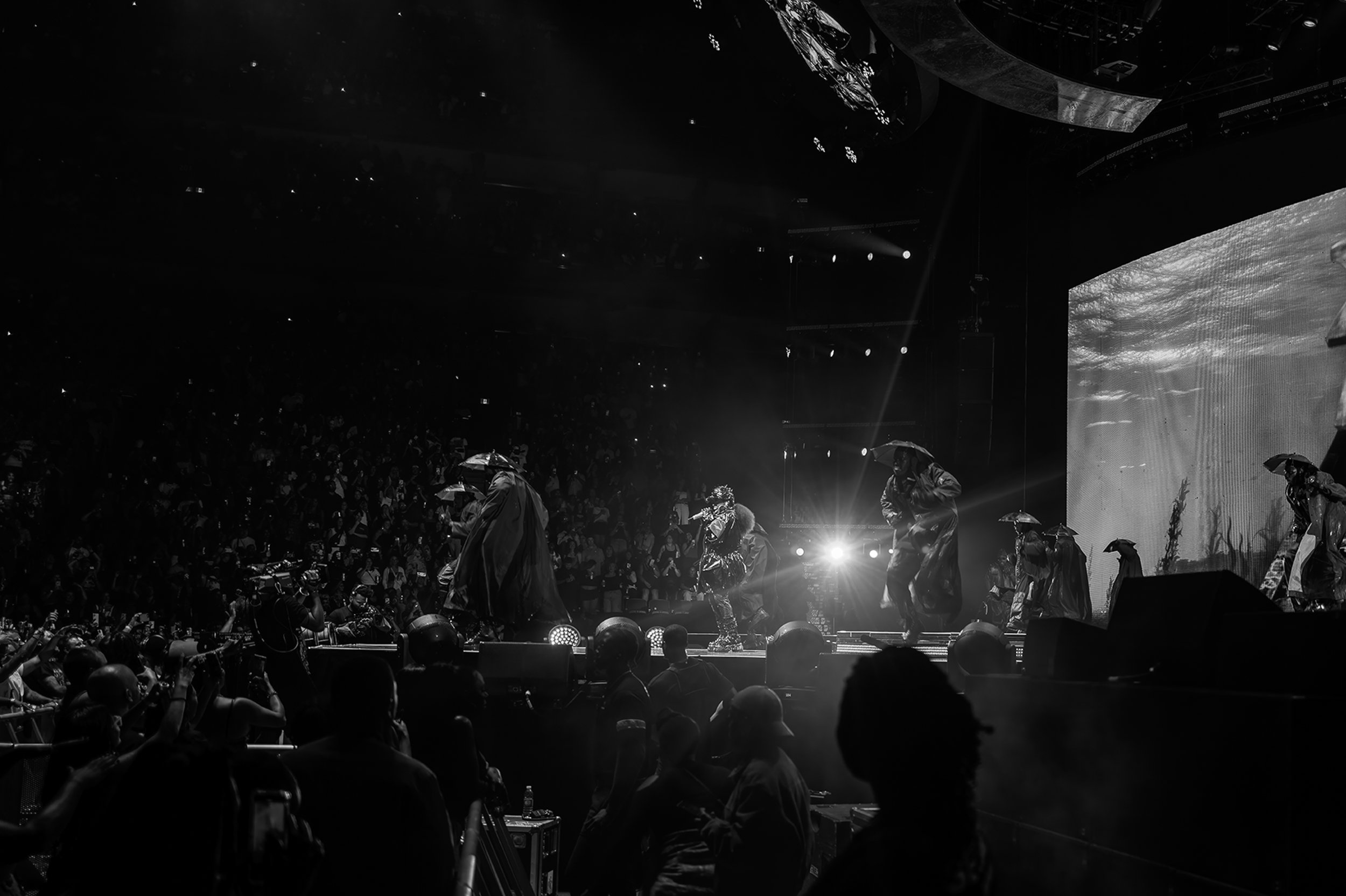 A live concert with performers on stage dressed in raincoats and holding umbrellas, with an audience watching in a darkened venue. The stage is illuminated by lights and a large video screen in the background displays visuals.