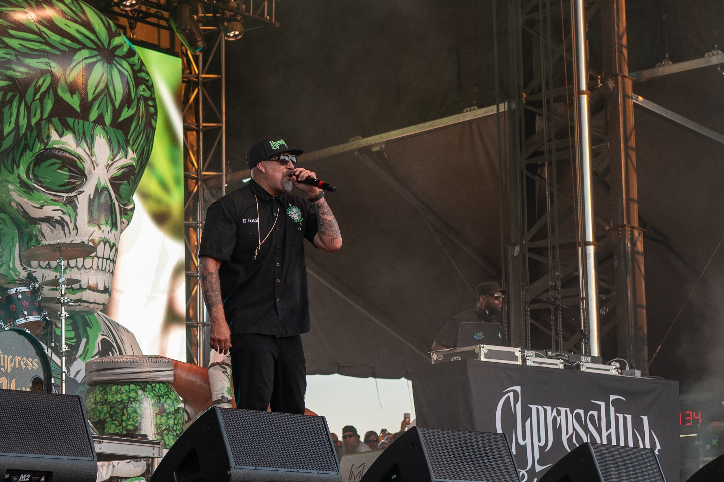 Musician with tattoos and sunglasses singing into a microphone on stage at a music festival, with a large backdrop featuring a green skull with pink hair and a drum on the stage.