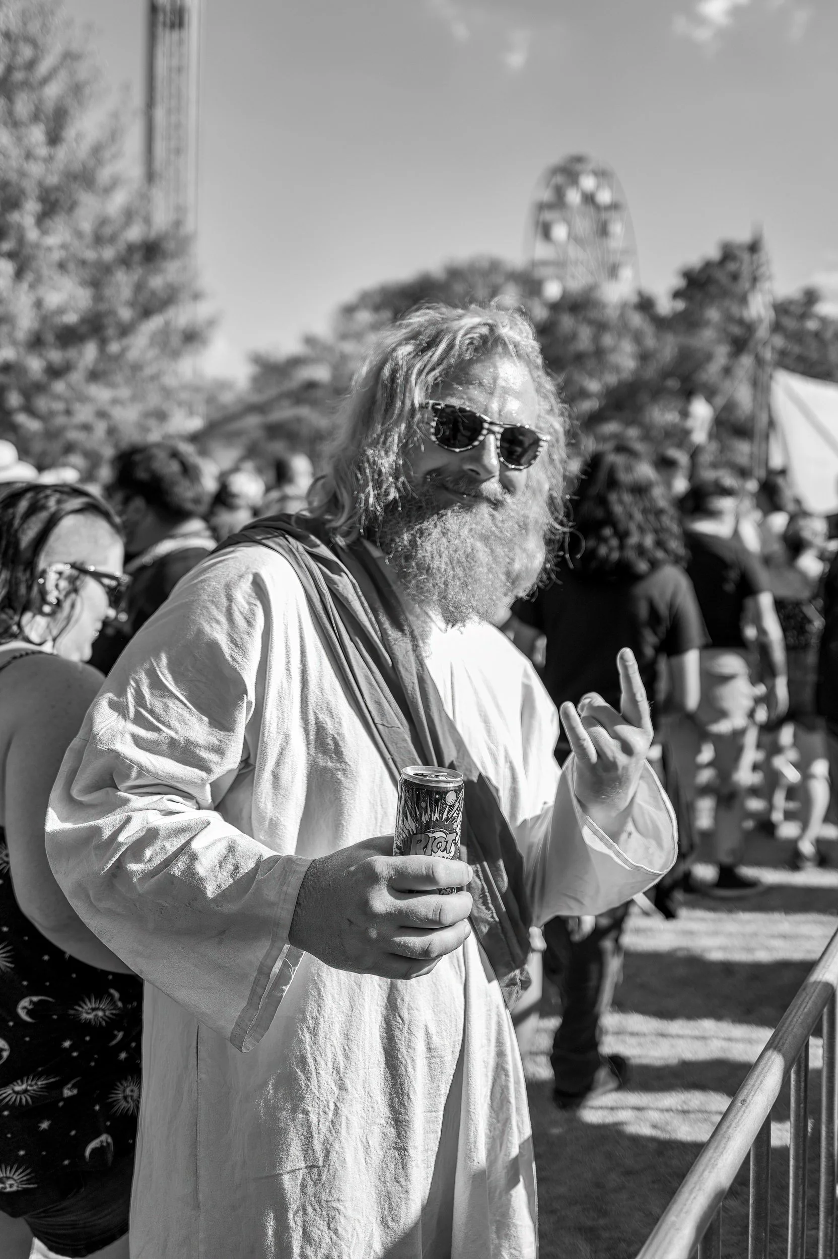 A bearded man with sunglasses holding a soda can at an outdoor event, making a peace sign with his other hand, with a crowd in the background.