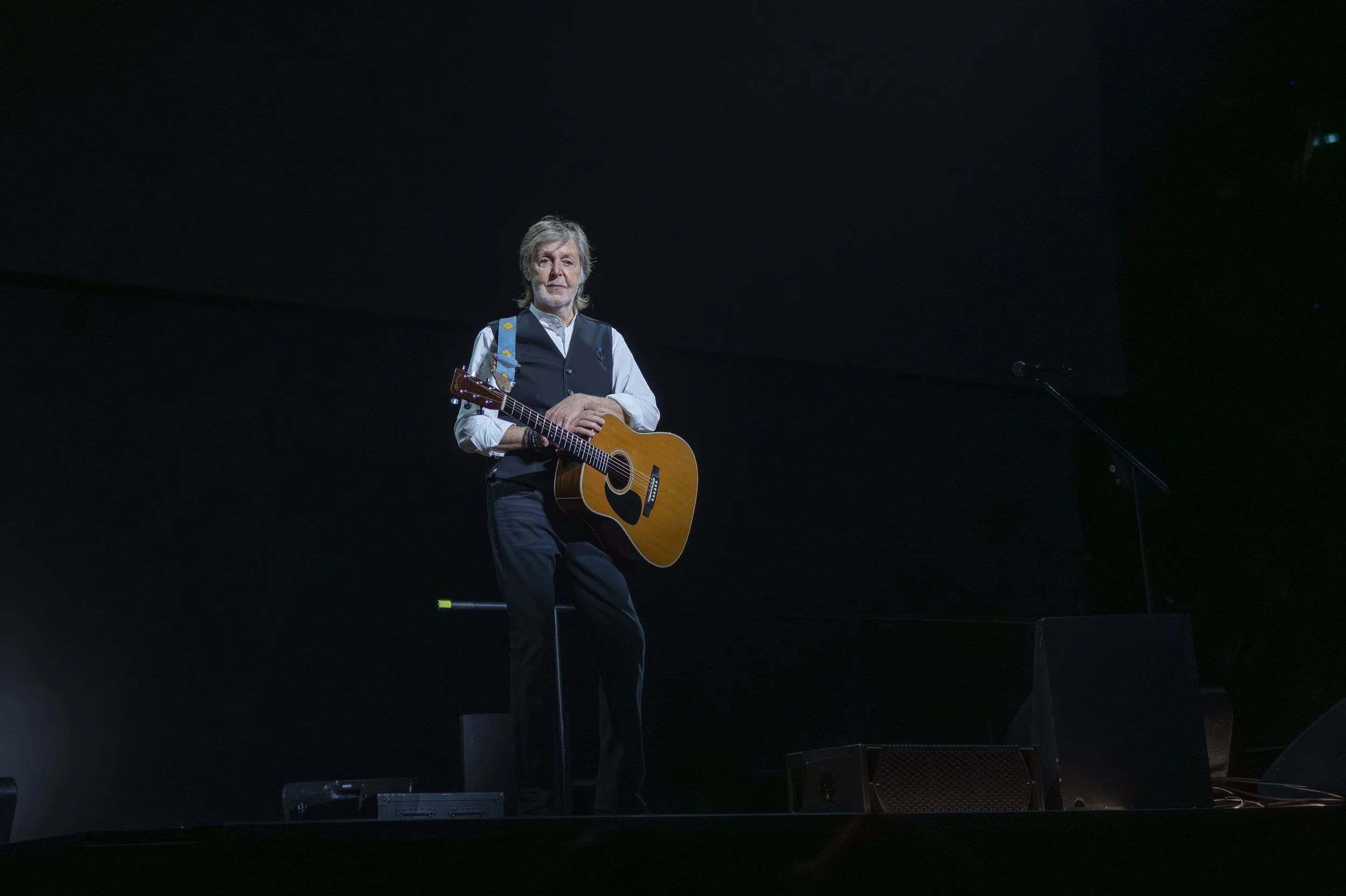 A man with gray hair and beard sitting on a stool on a dark stage, holding an acoustic guitar, with a microphone stand nearby.