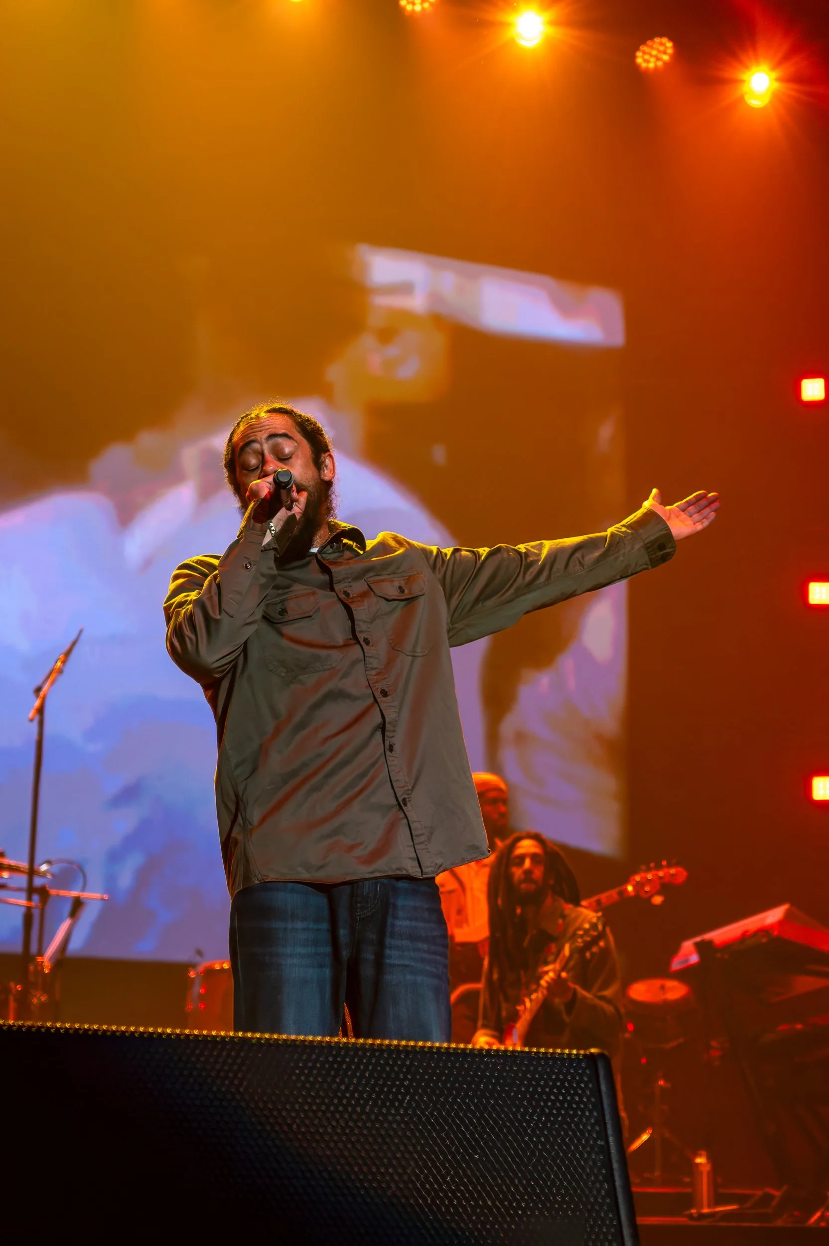 Performer singing with eyes closed on stage, surrounded by warm lighting and musical equipment, with large screen behind him displaying same scene.