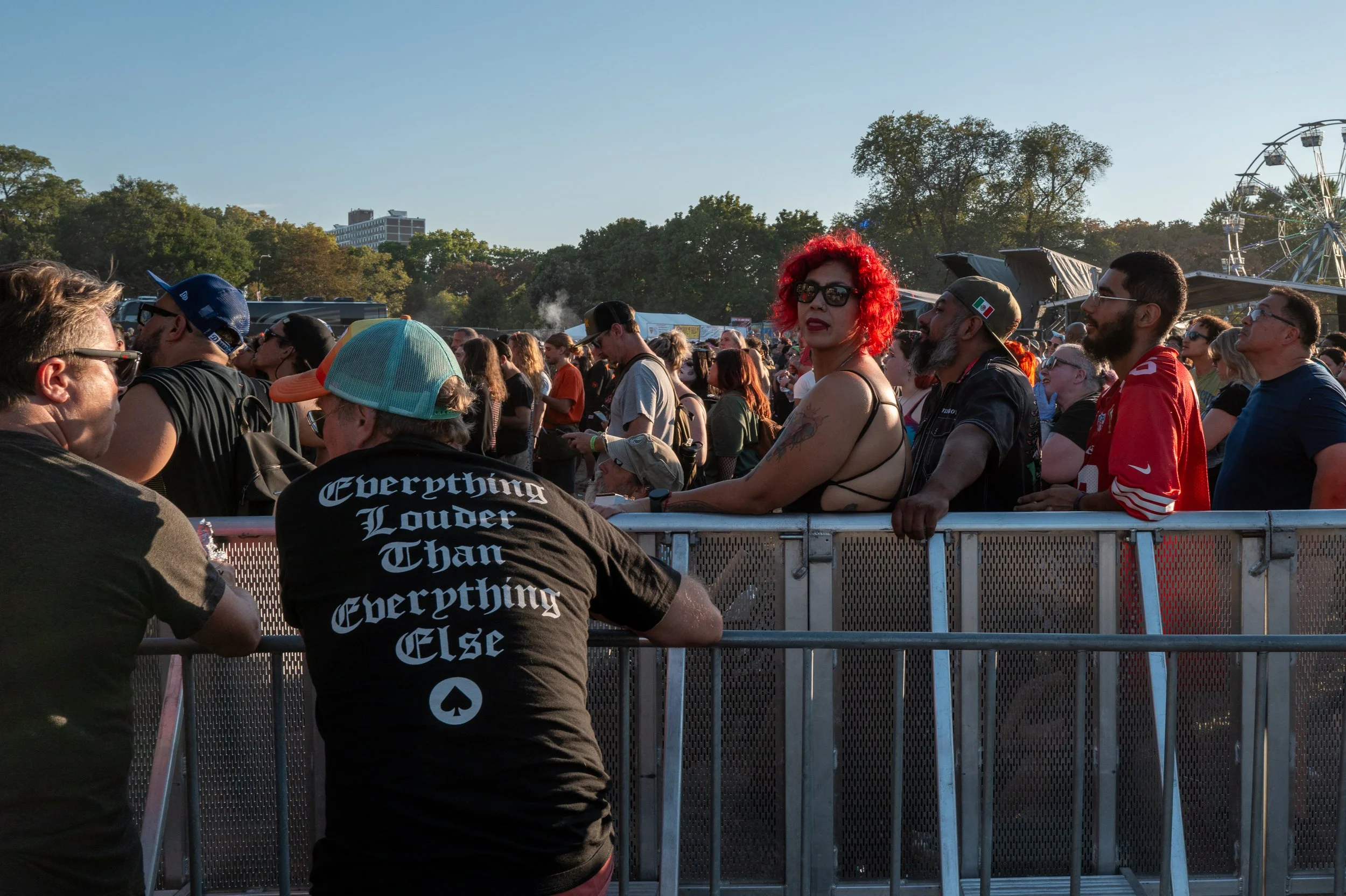Crowd of people attending an outdoor music festival, with some standing behind a metal barrier, and a Ferris wheel visible in the background.