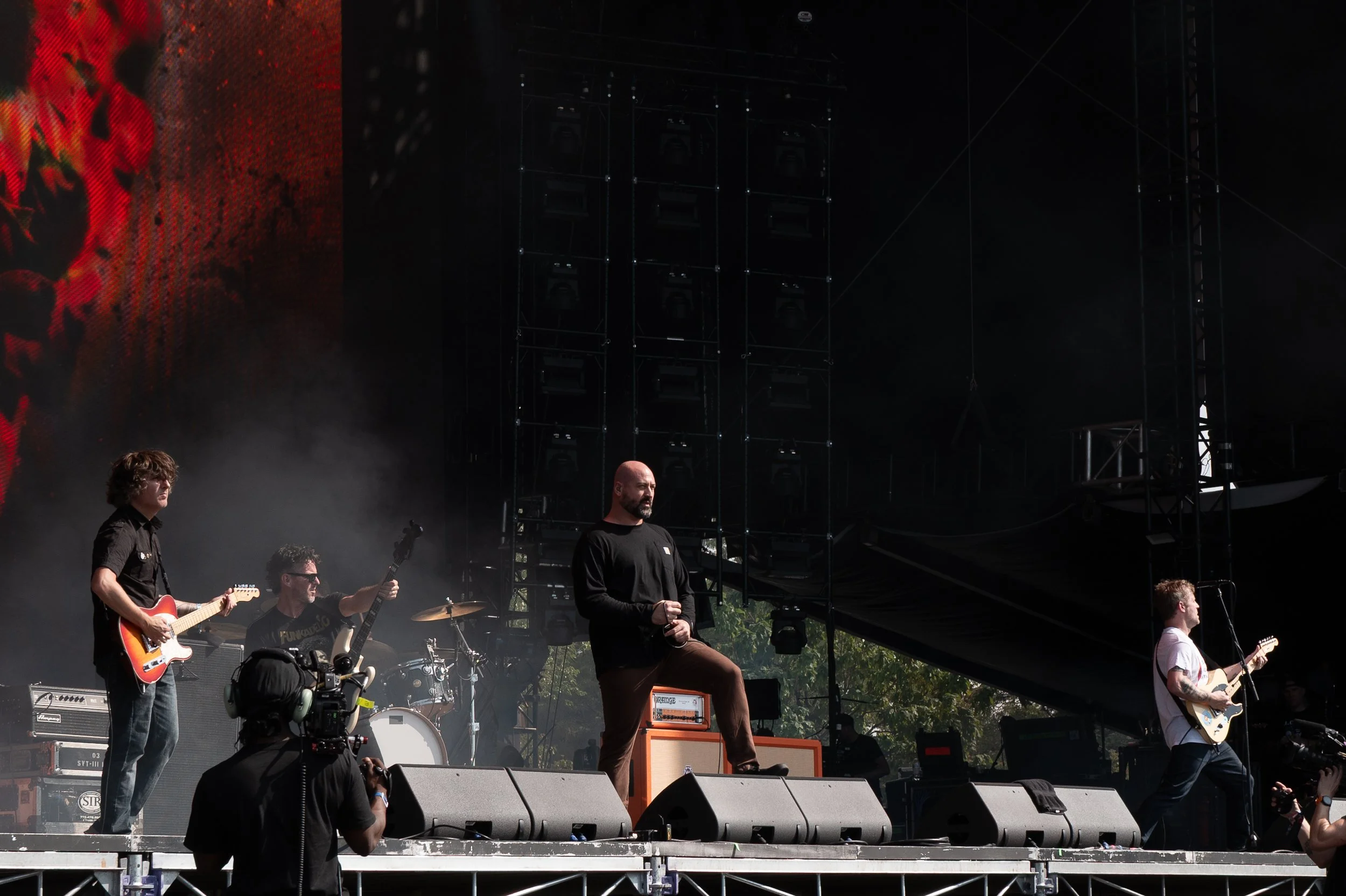 A band performing on an outdoor stage with four musicians playing guitars and drums.