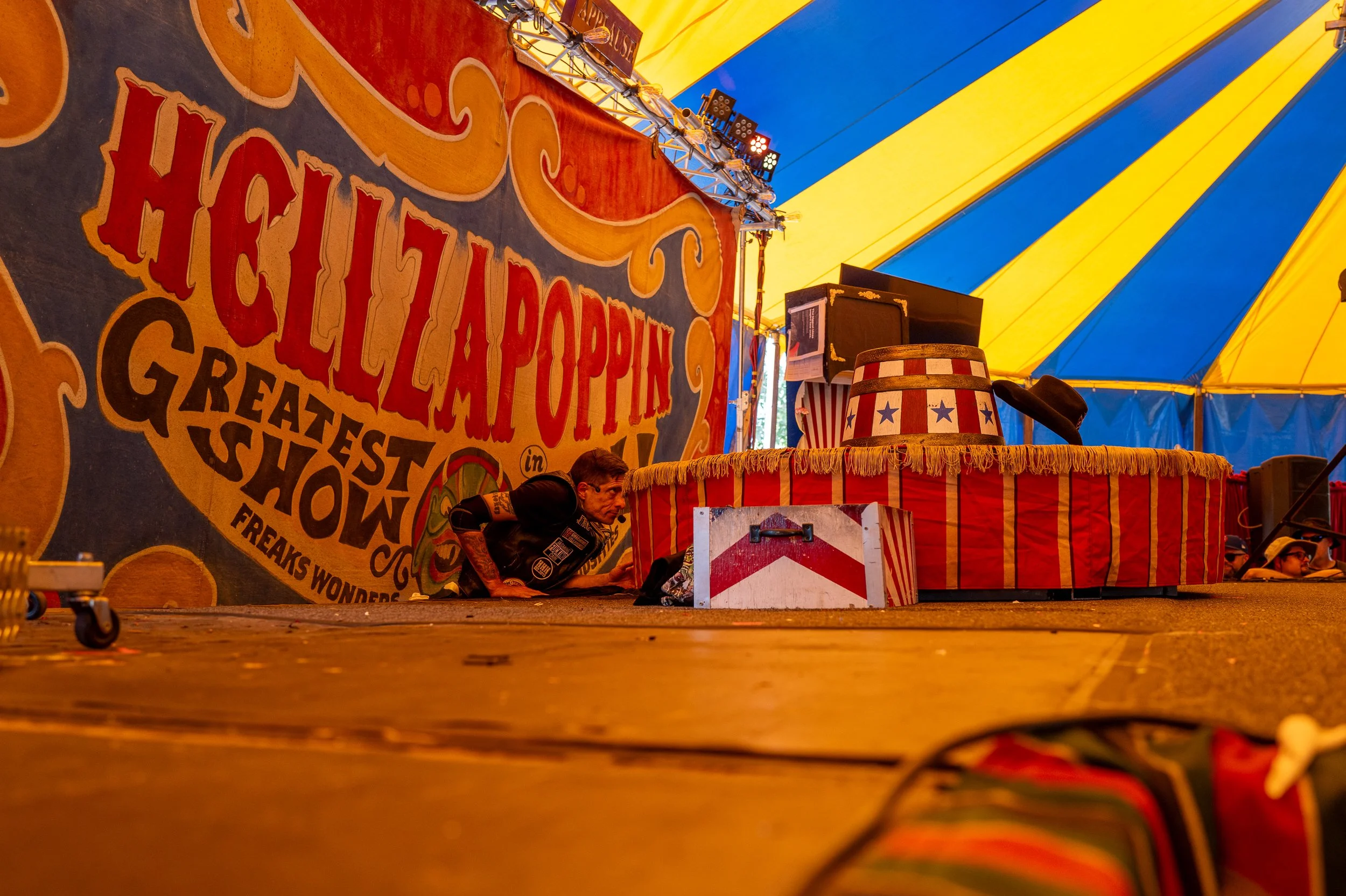 A performer crouches behind a circus ring, preparing for a show inside a colorful tent with a yellow and blue canopy. A large sign reads 'Heidi LaPoppin Greatest Show in Freaks Wonders.' There are stage props, including a hat and a drum decorated wit