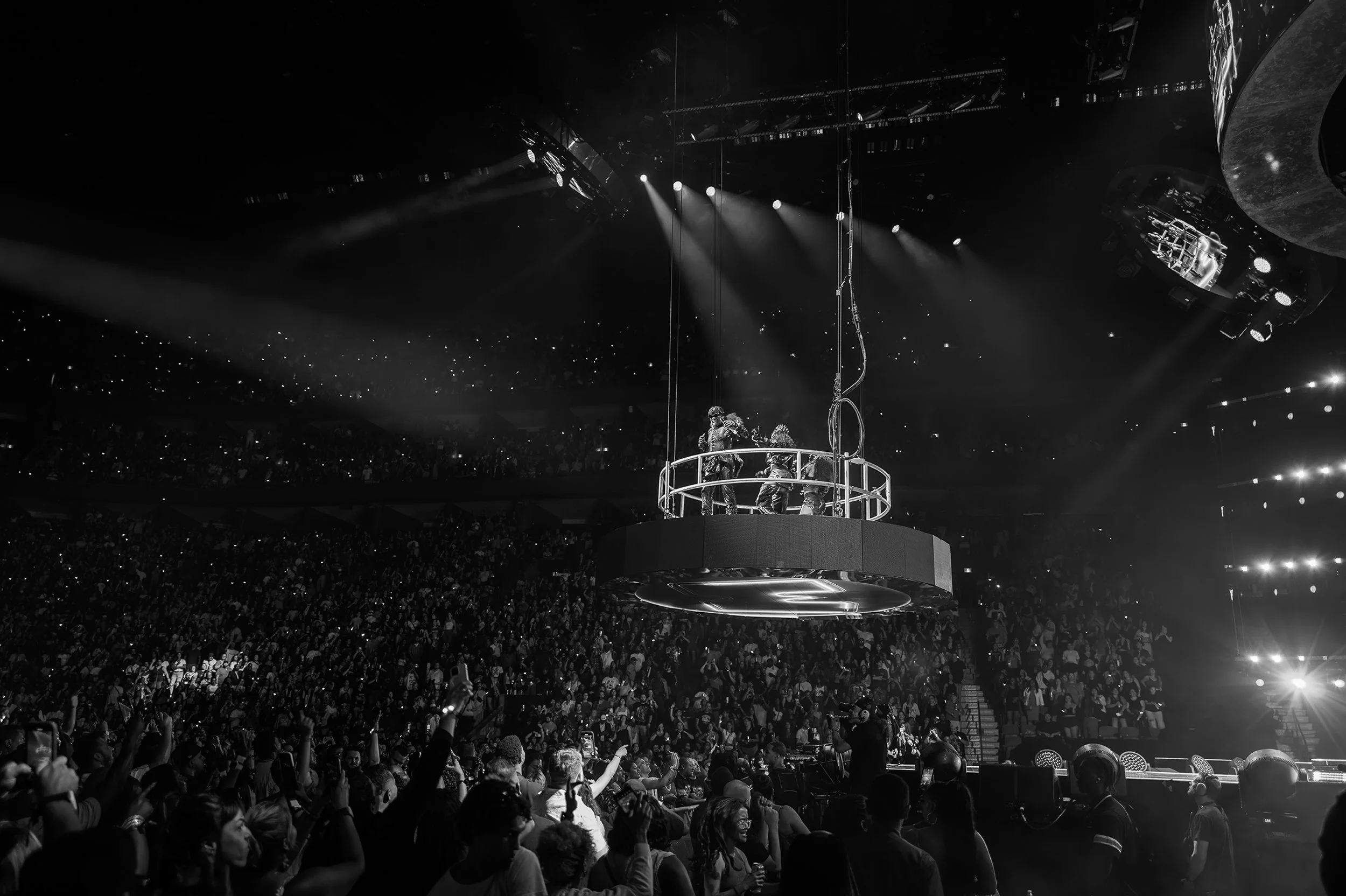 Black and white photo of a live concert with performers on a suspended circular platform surrounded by a large crowd in an indoor arena.