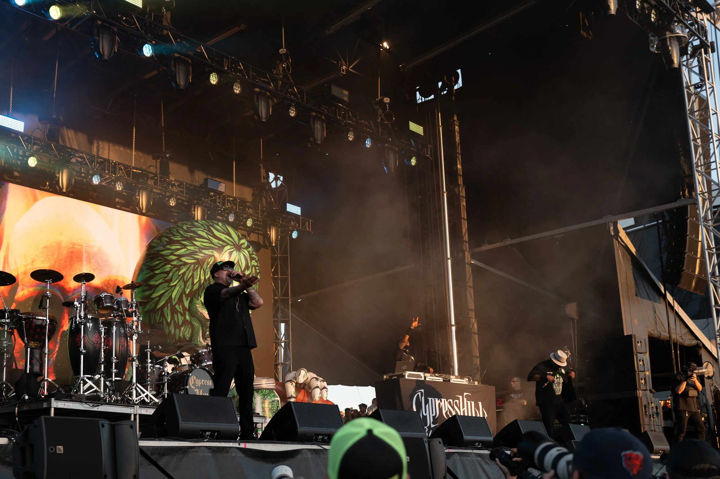 Musicians performing on an outdoor stage with a large colorful backdrop, including a tiger design, various musical instruments, and sound equipment. Audience members are visible in the foreground.