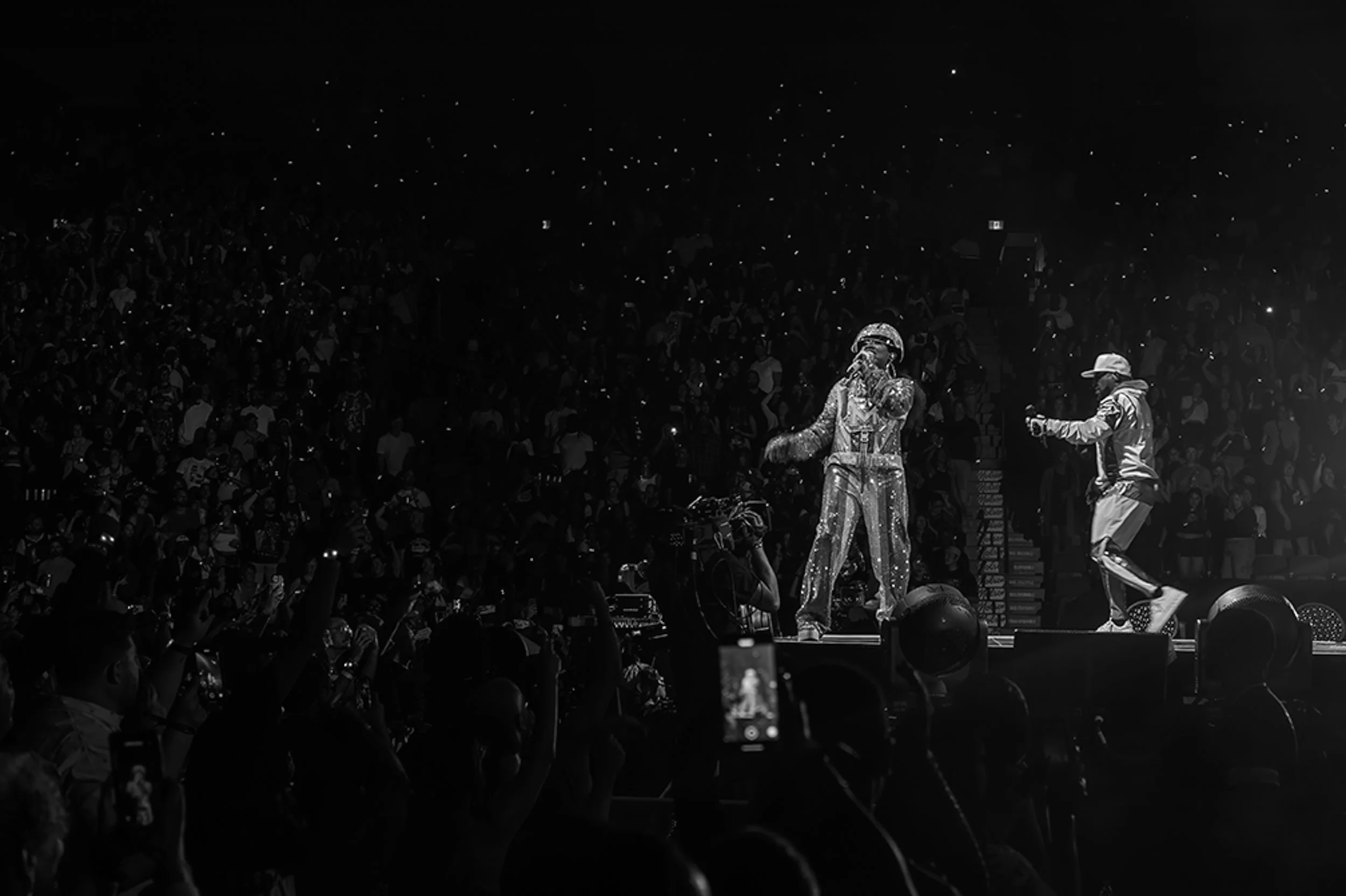 A performer dressed in a sparkling, elaborate outfit on stage, with another person in casual clothing near him, during a concert or event with a large crowd watching.