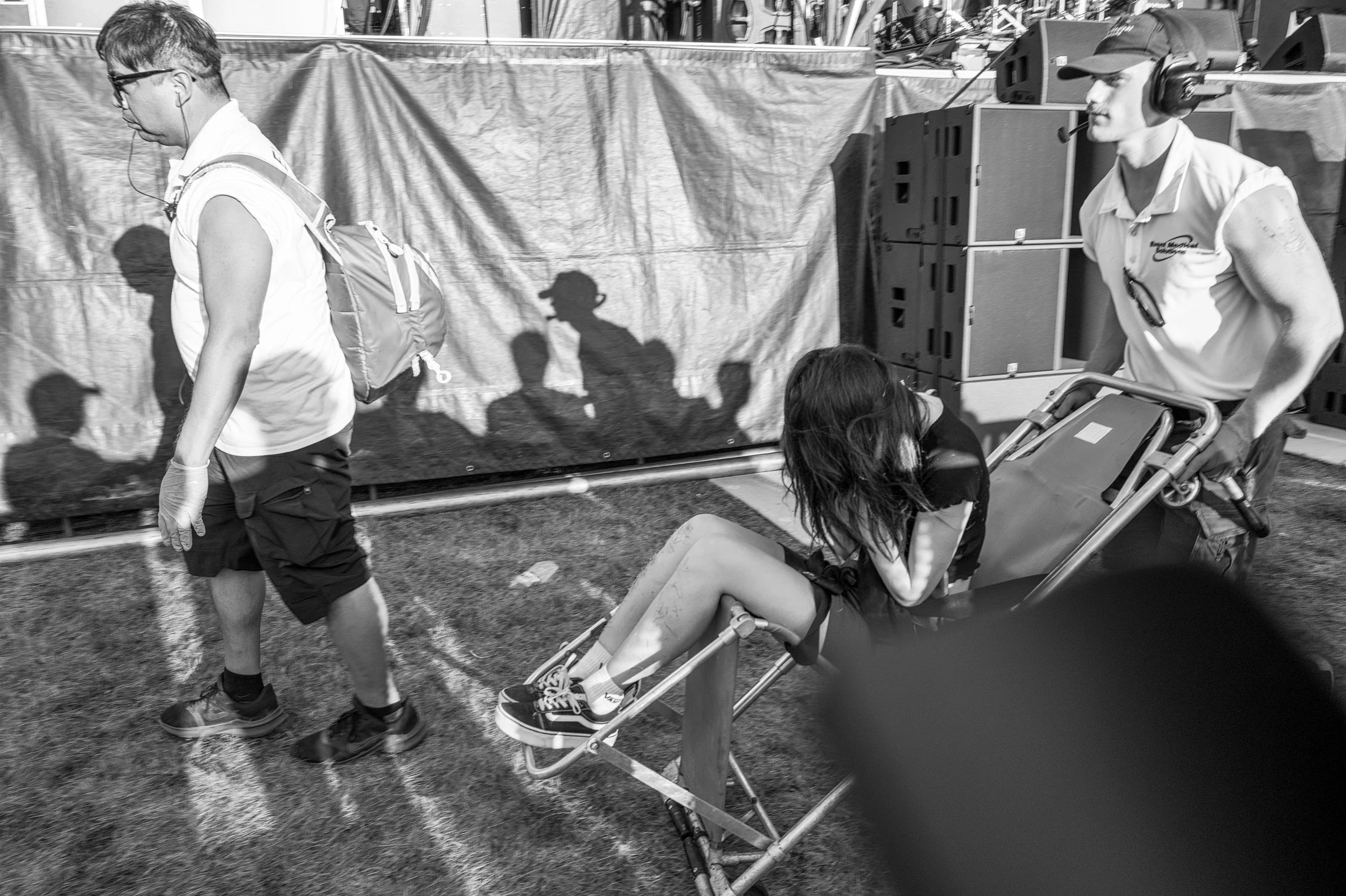 A woman is sitting on a stretcher being pushed by a man at an outdoor event, with a third man walking nearby. The woman is wearing headphones and casual clothing, and the men are in casual athletic attire. Shadows of people are cast on a tent or back