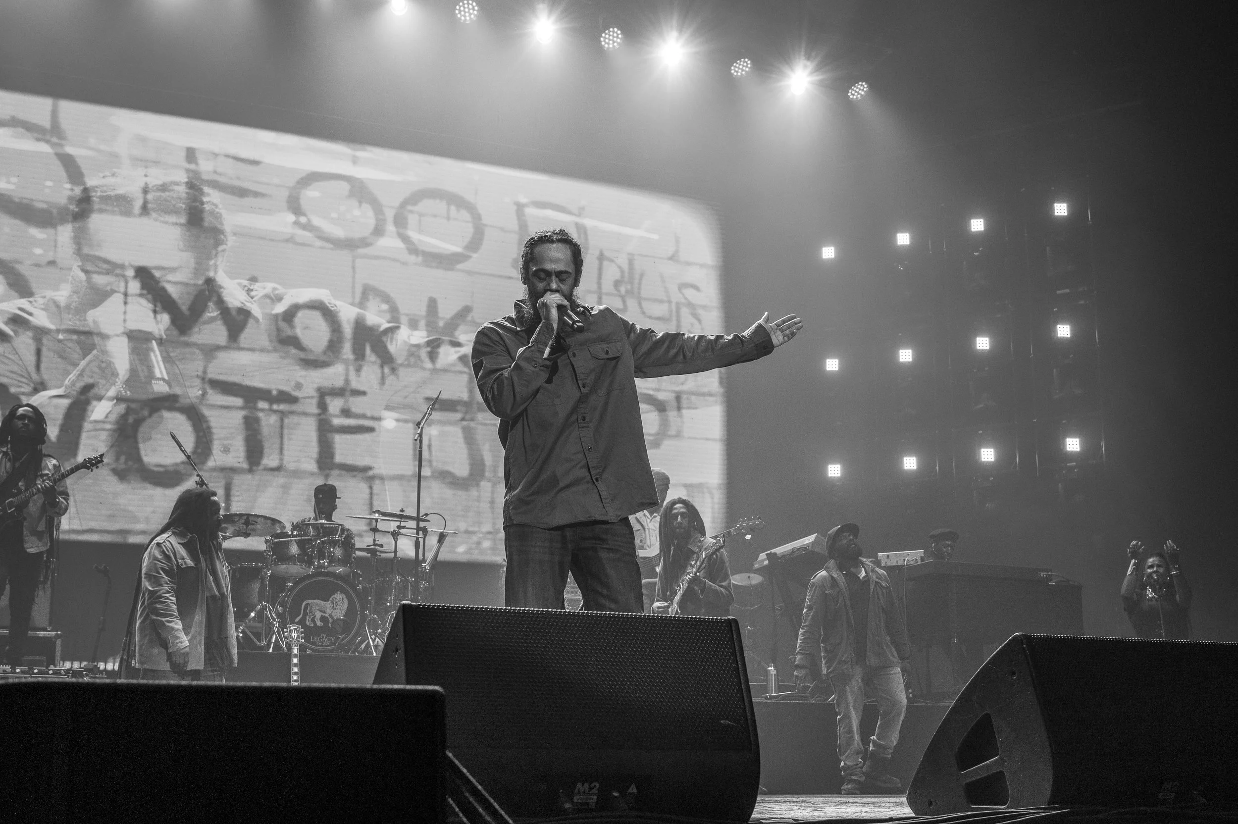 A male singer performs on stage with a microphone, accompanied by a band, in a concert setting with bright lights and a large screen in the background.