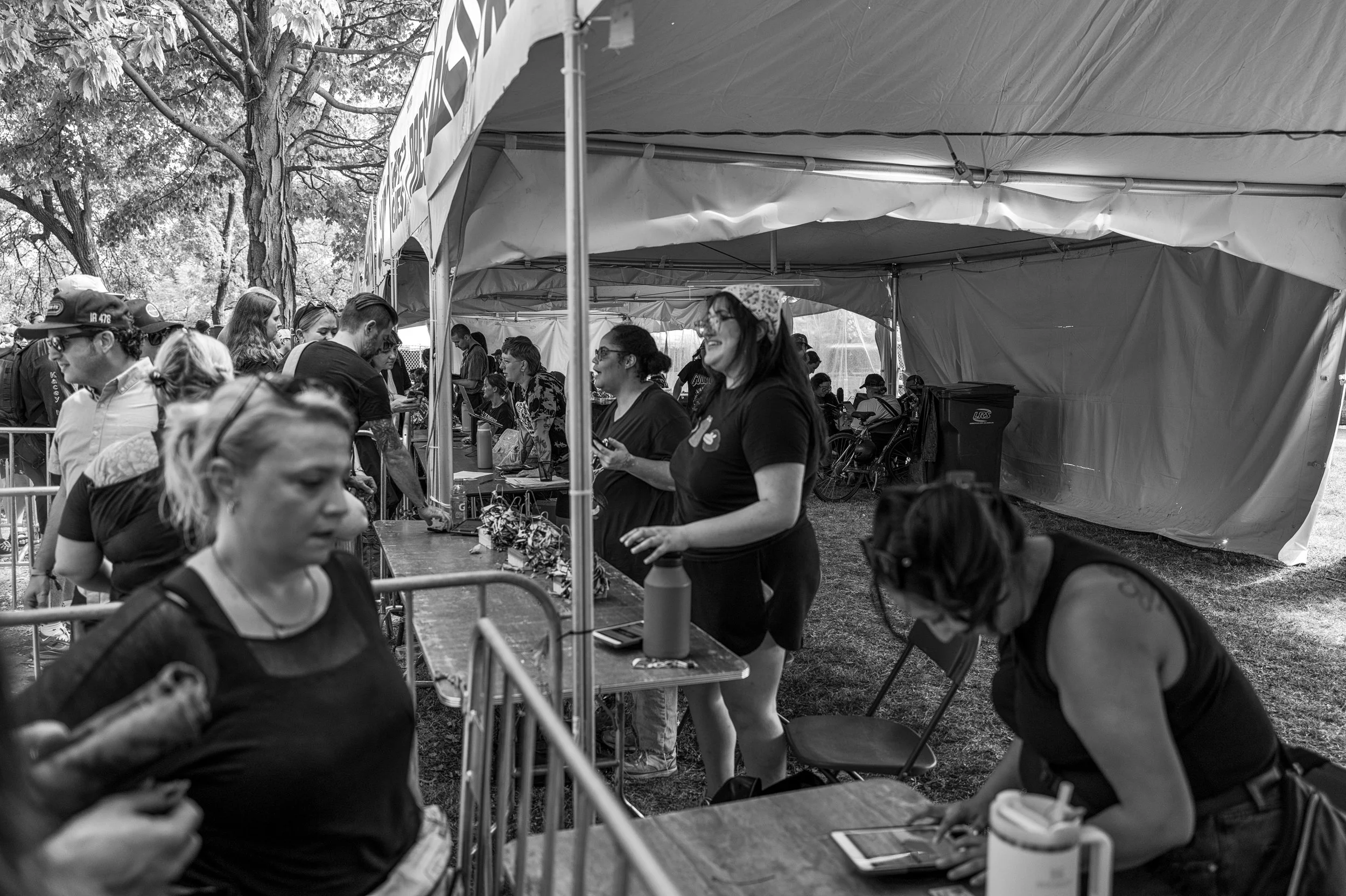 A black and white photo of people at an outdoor event under a large tent, with a long table where several women are serving or managing items, and others waiting in line or walking by. The event appears lively with trees visible in the background.