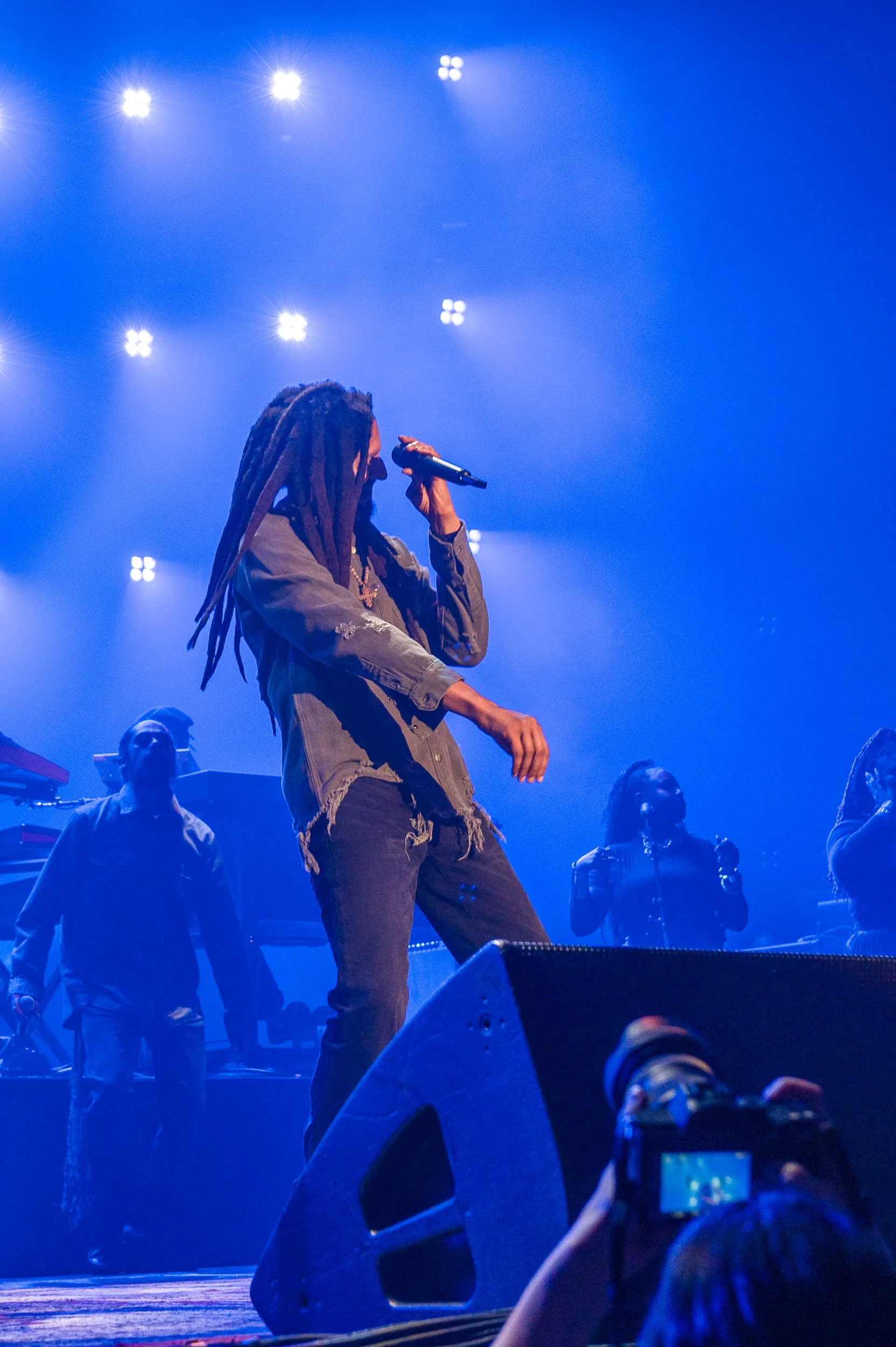 Performer with dreadlocks singing into a microphone on stage illuminated by blue lights, with backup singers and musicians in the background.