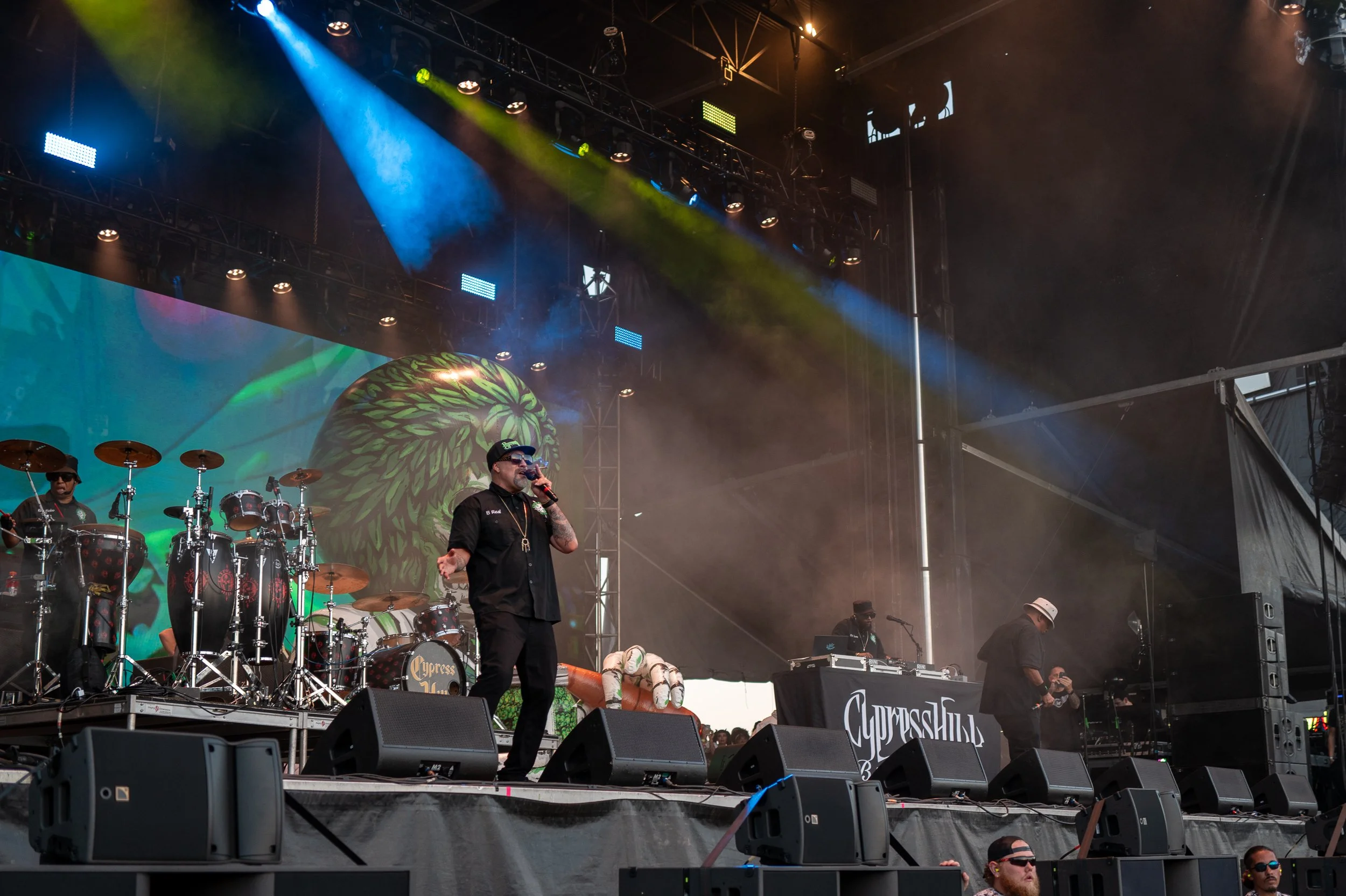 Stage with band performing at Cypress Hill concert, featuring a drummer, a singer with sunglasses and hat, DJ, and large animated backdrop.