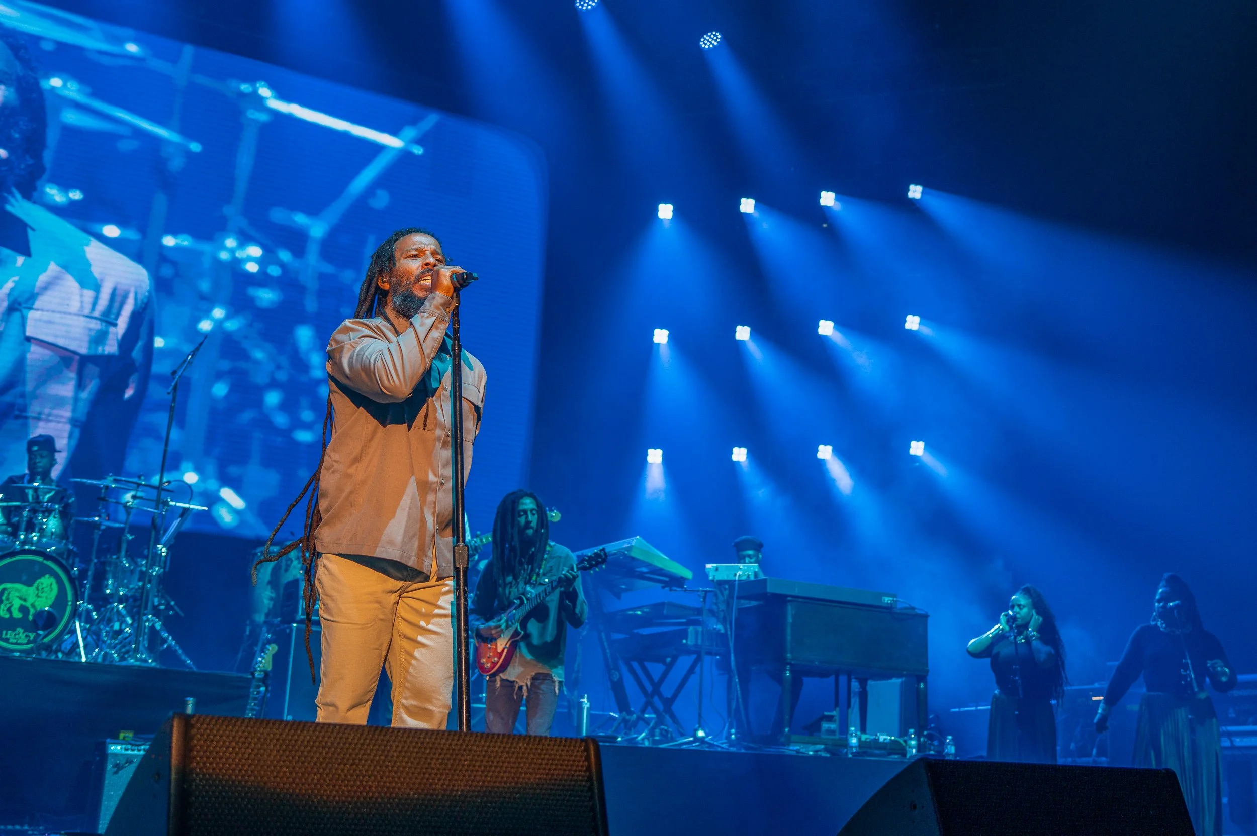 A band performs on stage with blue lighting, featuring a singer with dreadlocks holding a microphone, other band members playing instruments, and in the background a large screen showing a live video feed of the performance.