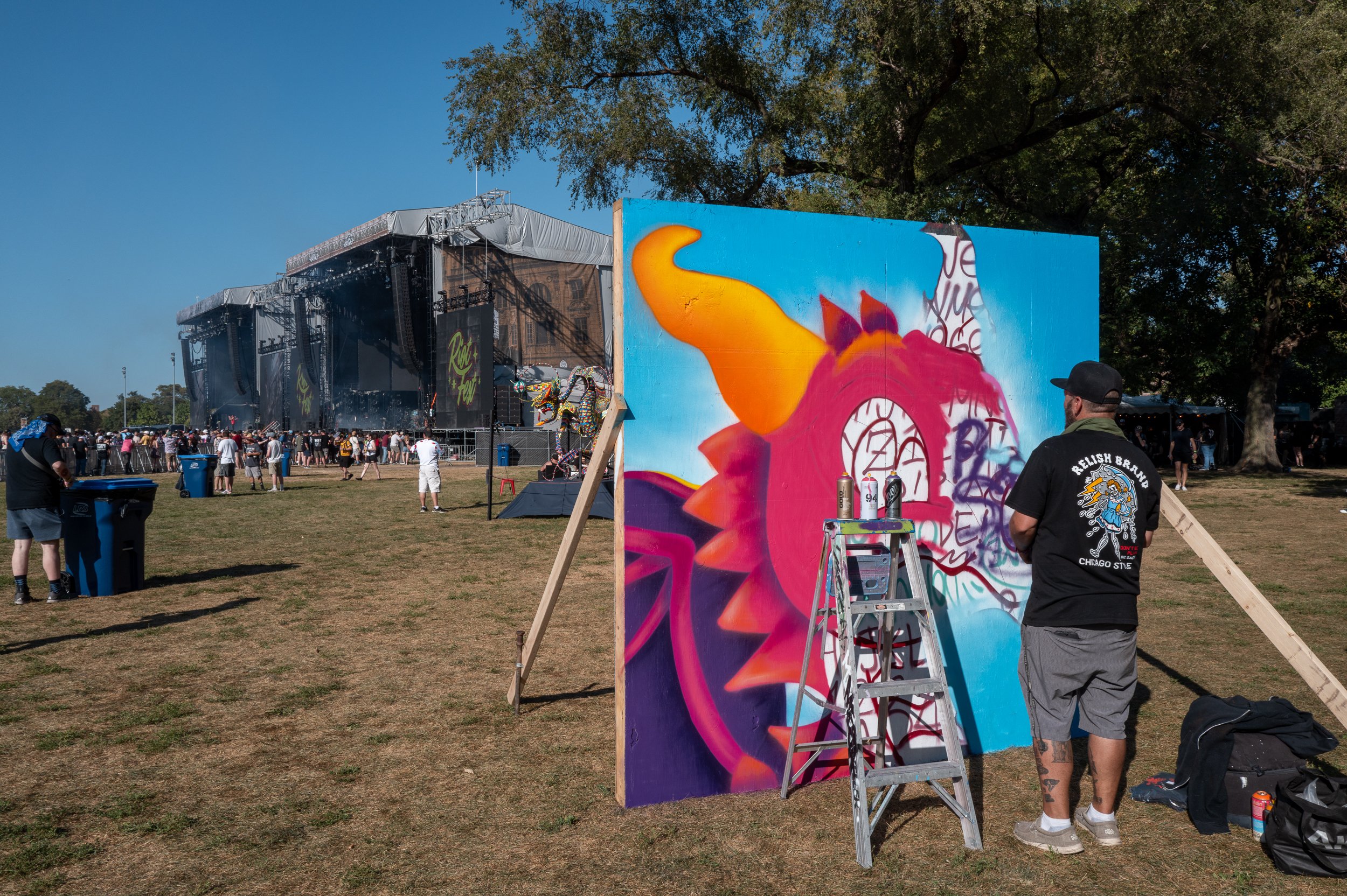 Artist painting graffiti art on a large board at an outdoor music festival with a stage in the background and attendees walking around.