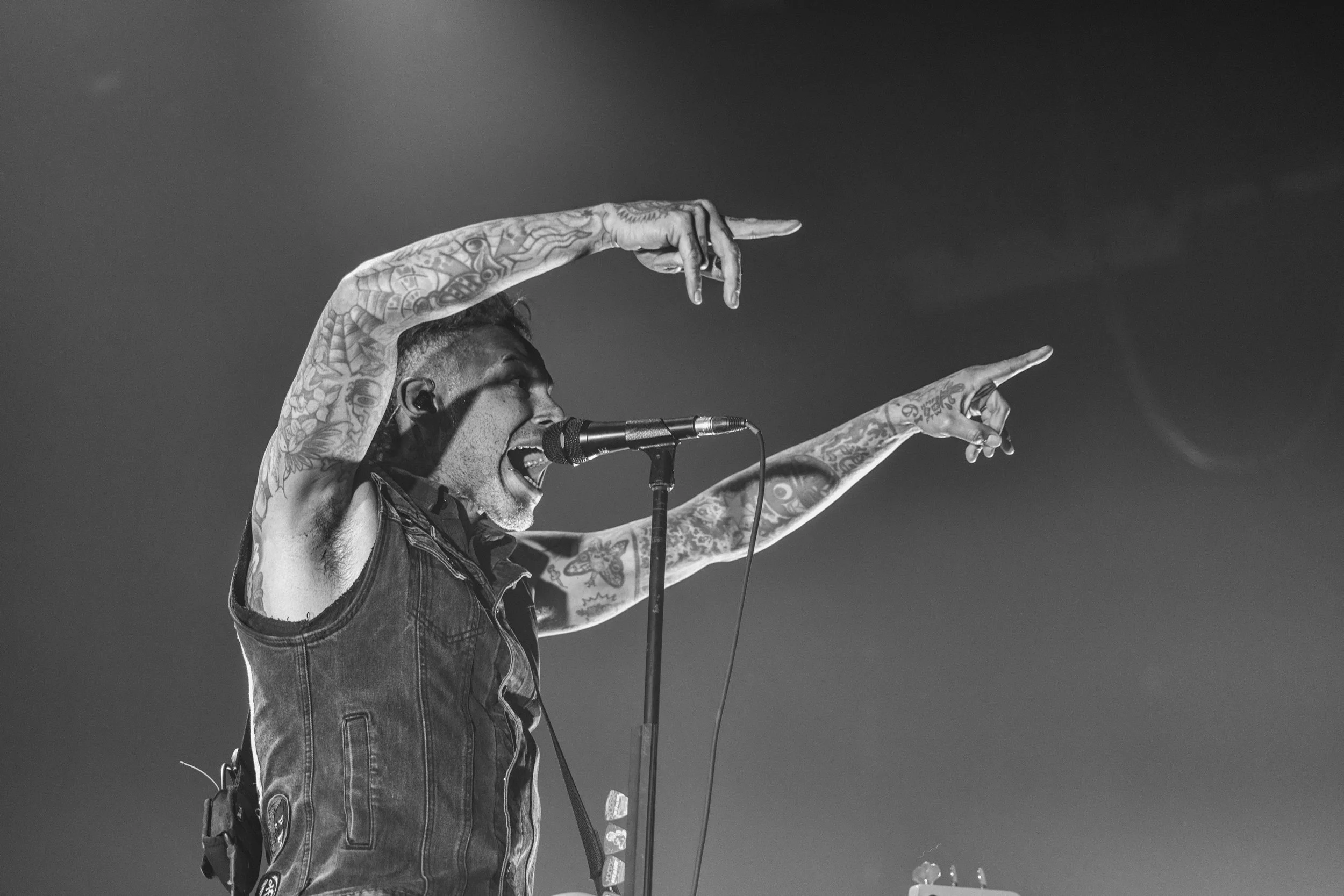 A tattooed male singer passionately singing into a microphone on stage, with arms raised and fingers pointing.