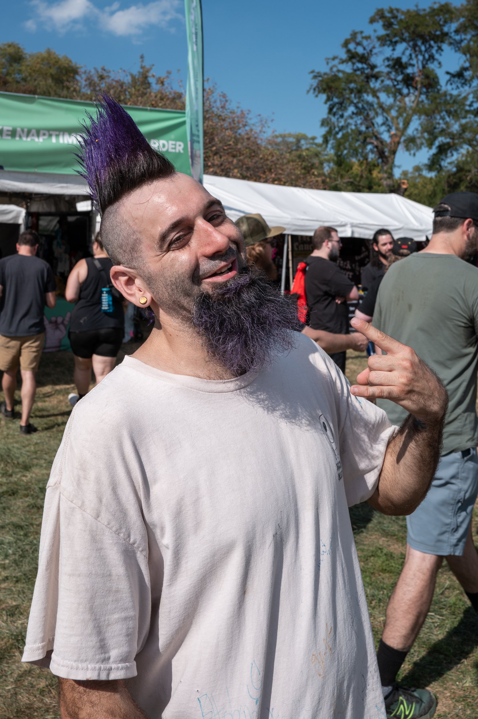 A man with a tall purple mohawk hairstyle and a purple beard, pointing to himself and smiling at an outdoor event with tents and other people.