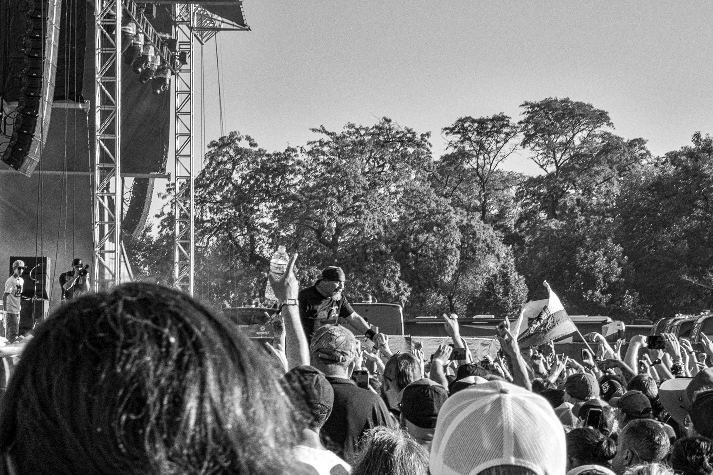 Black and white photo of a large outdoor concert with a crowd, stage, and musicians playing, outdoors with trees in the background.