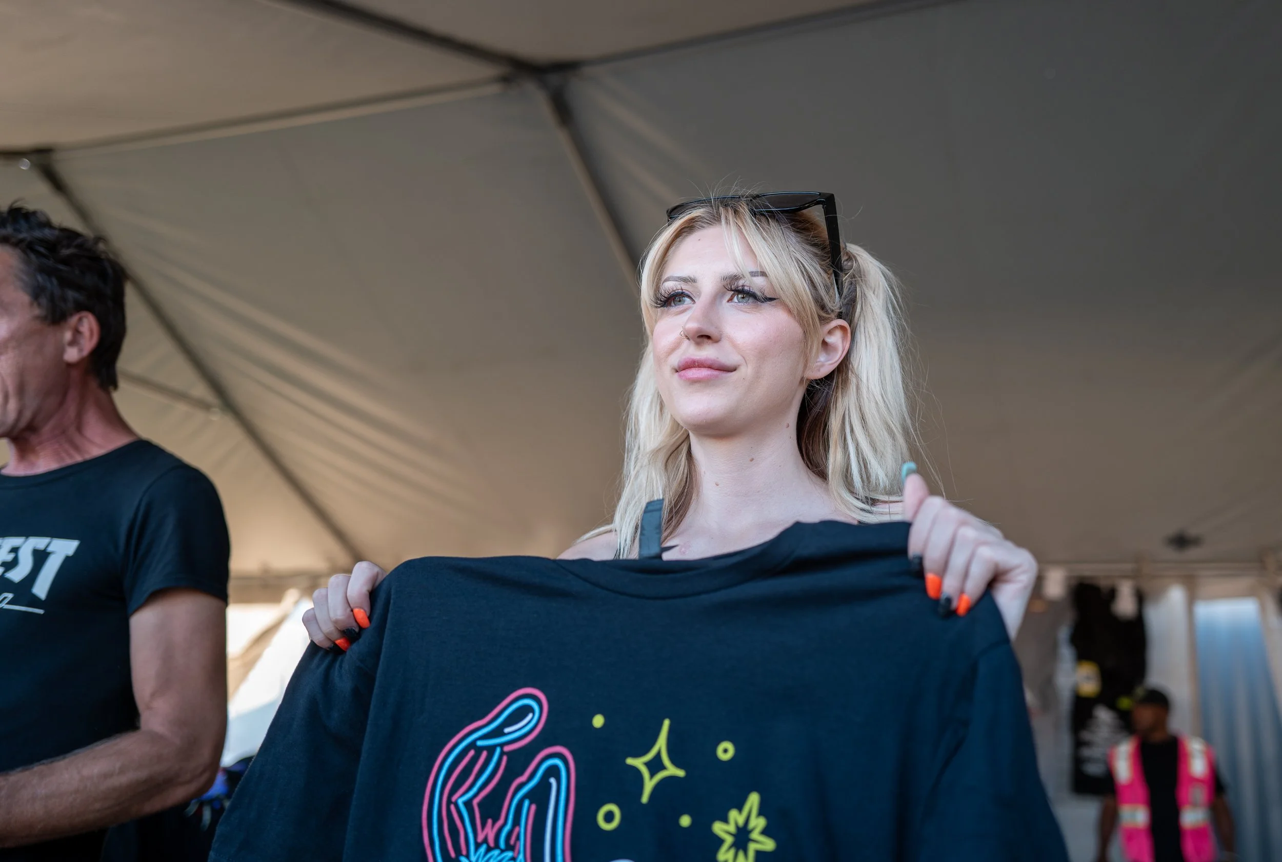 A young woman with blonde hair and glasses on her head holding up a black t-shirt with colorful neon-like designs at an outdoor event under a large tent.
