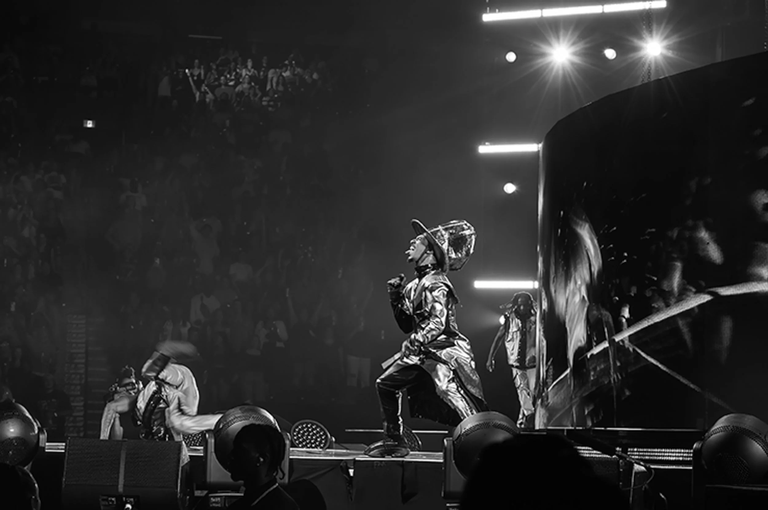 Performer singing on stage during concert, wearing metallic outfit and cowboy hat, with large face projection on screen behind, audience in background, bright stage lights.