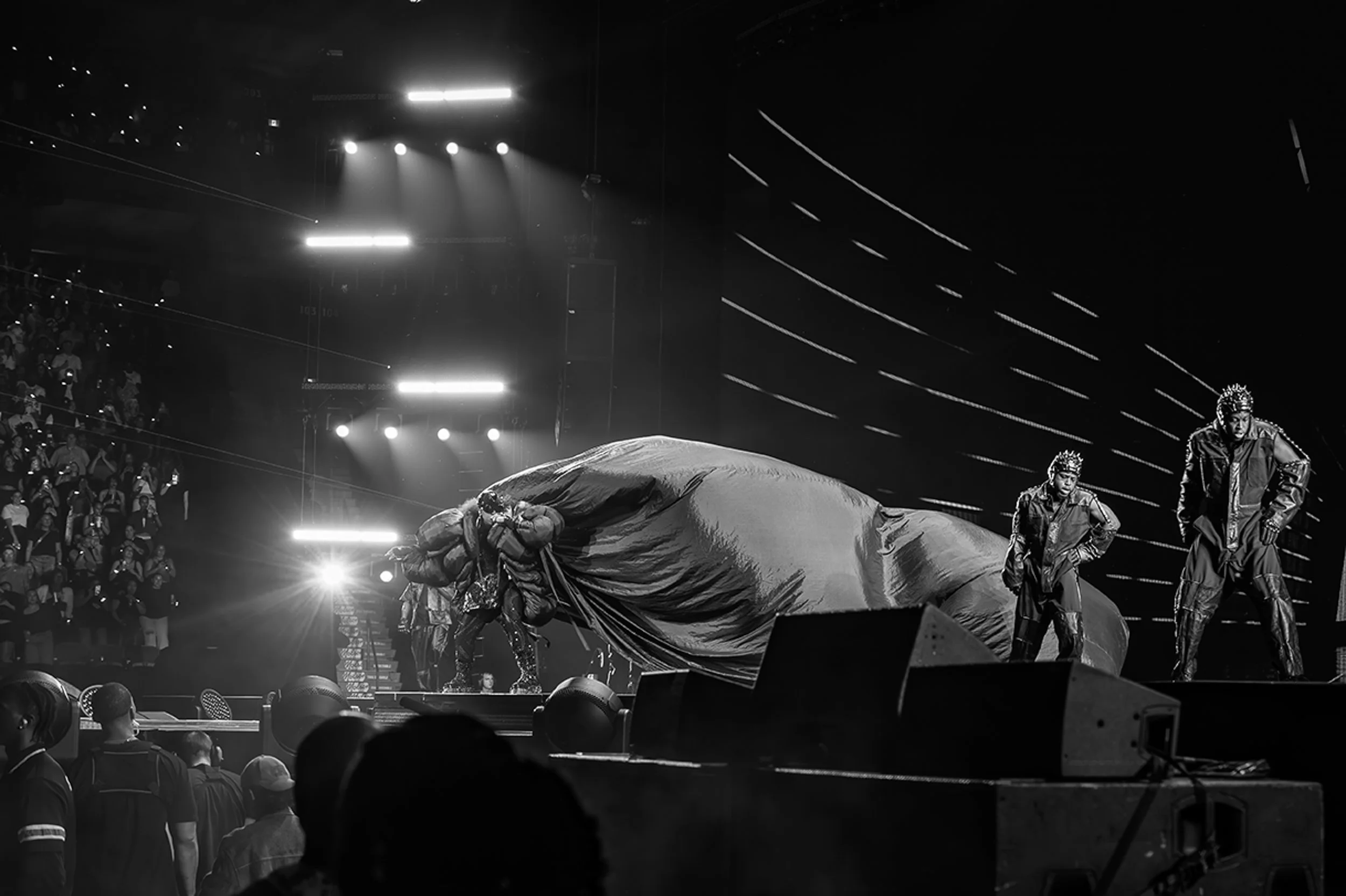 Performers on stage revealing a large object under a black cloth with dramatic lighting, audience in the background.